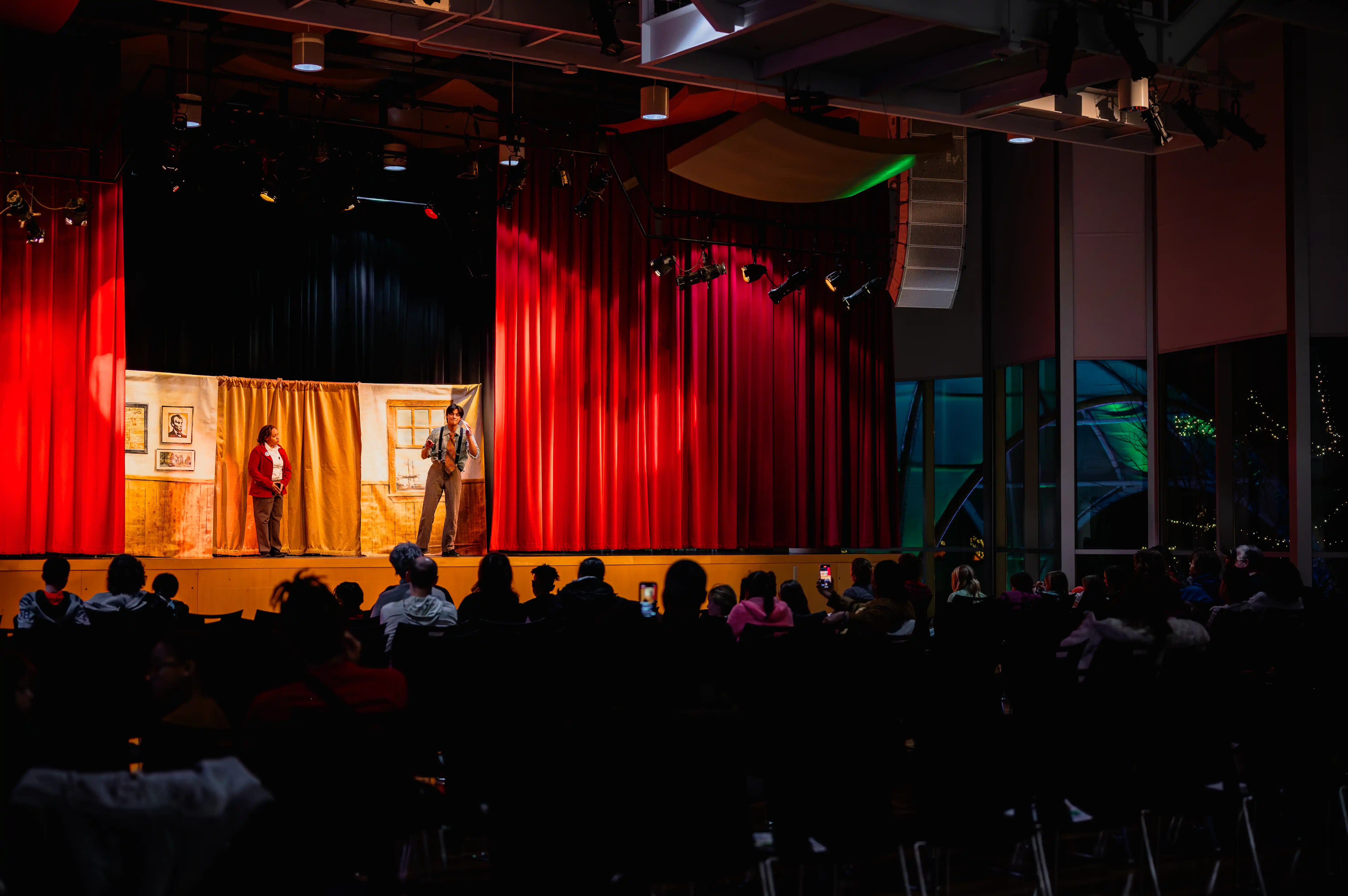 Audience viewing performers on a stage with red curtains at a dimly lit event.