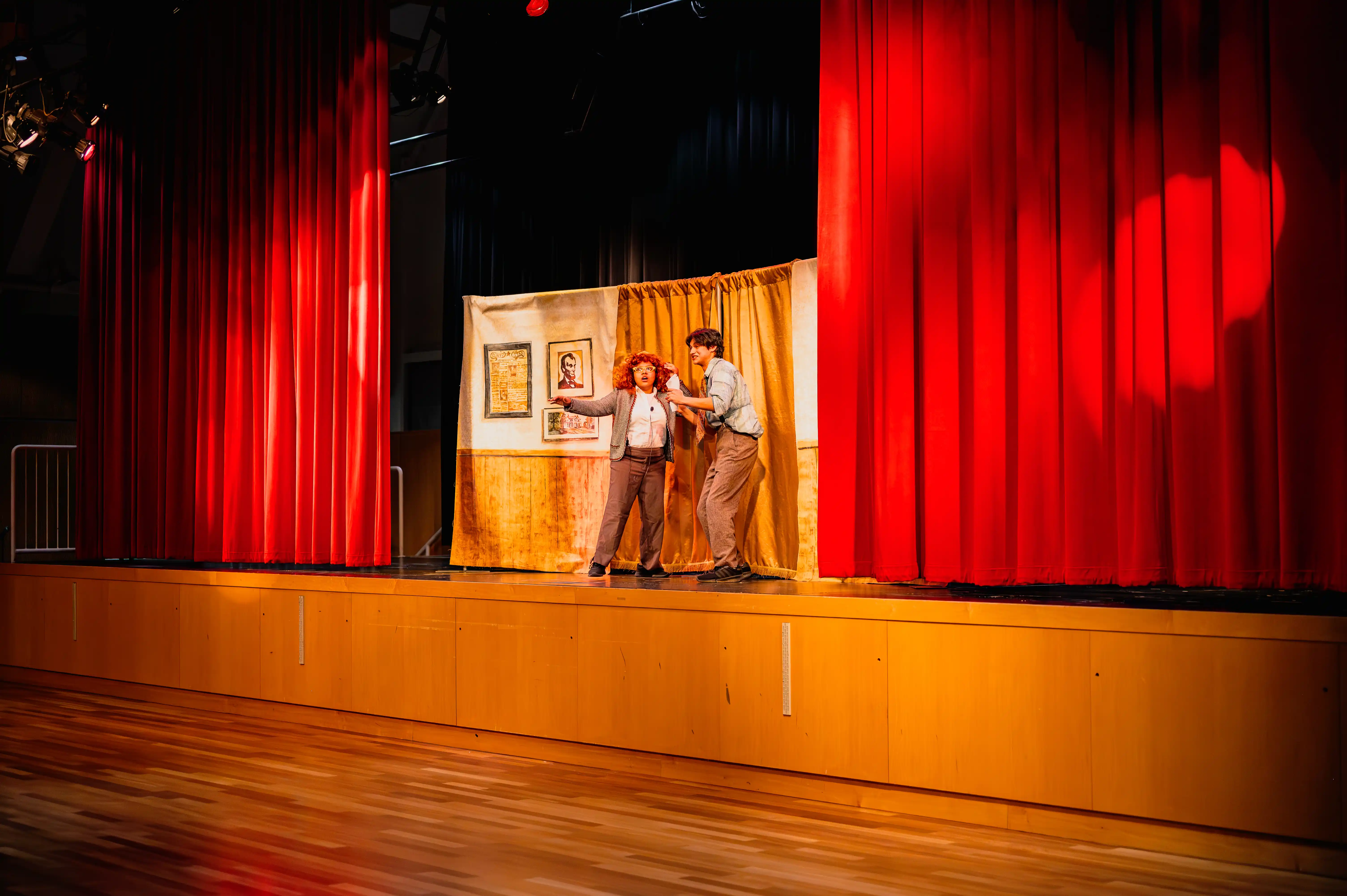 Two actors on stage during a theater performance with red curtains in the background.