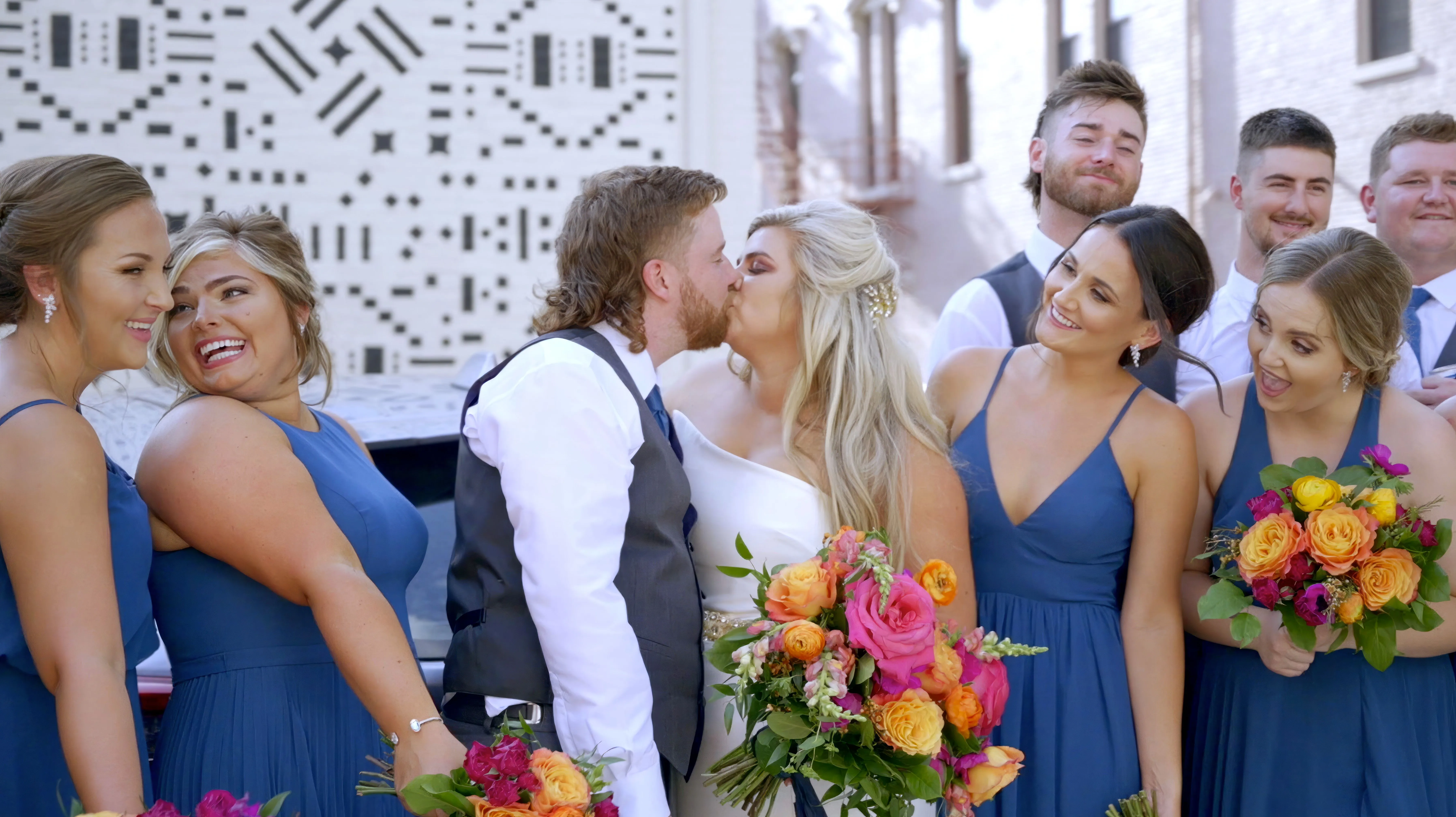 Wedding party with a couple kissing and bridesmaids in blue dresses and groomsmen smiling in the background.
