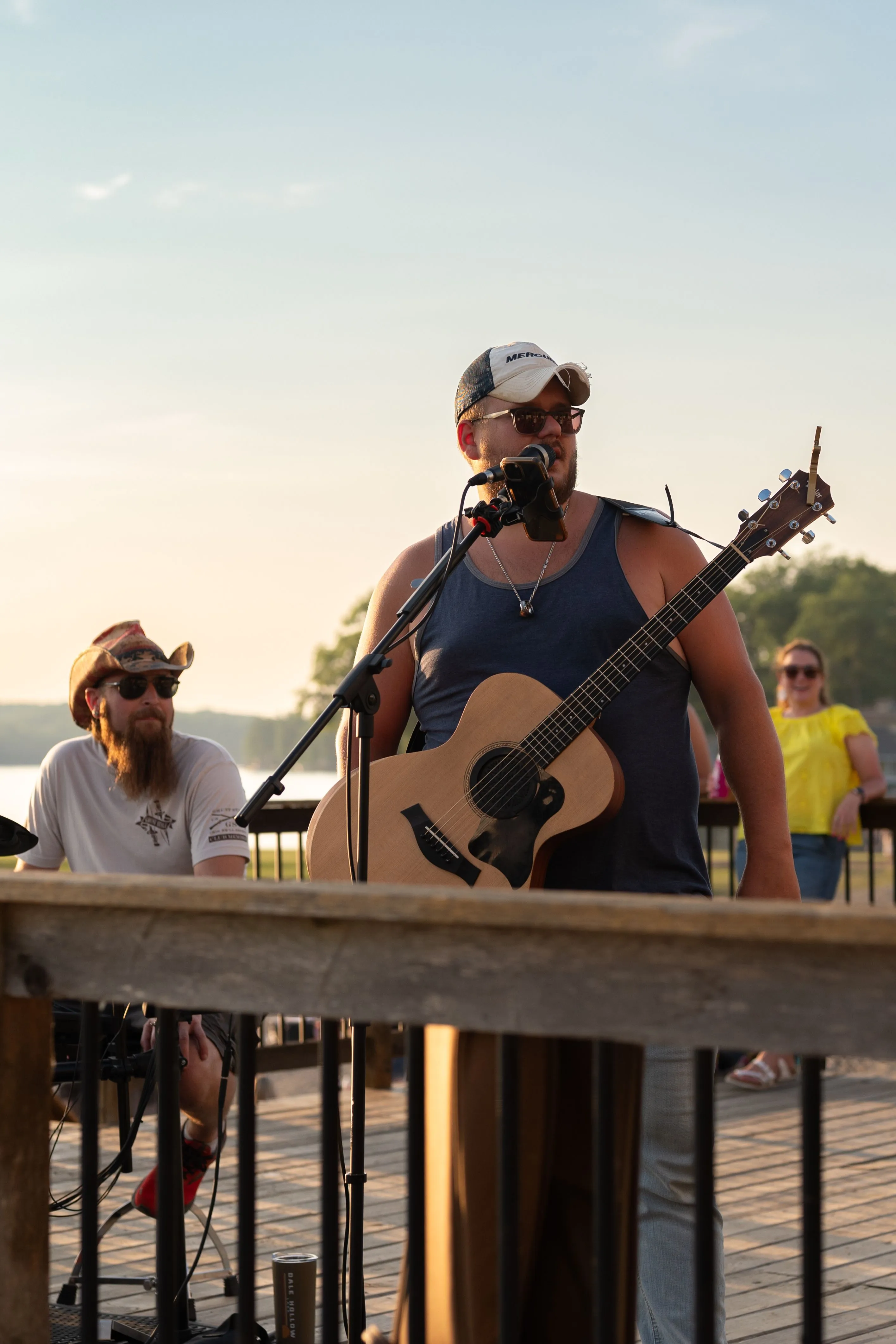 Musician playing guitar outdoors on a deck with another person in the background and a waterfront view during sunset.