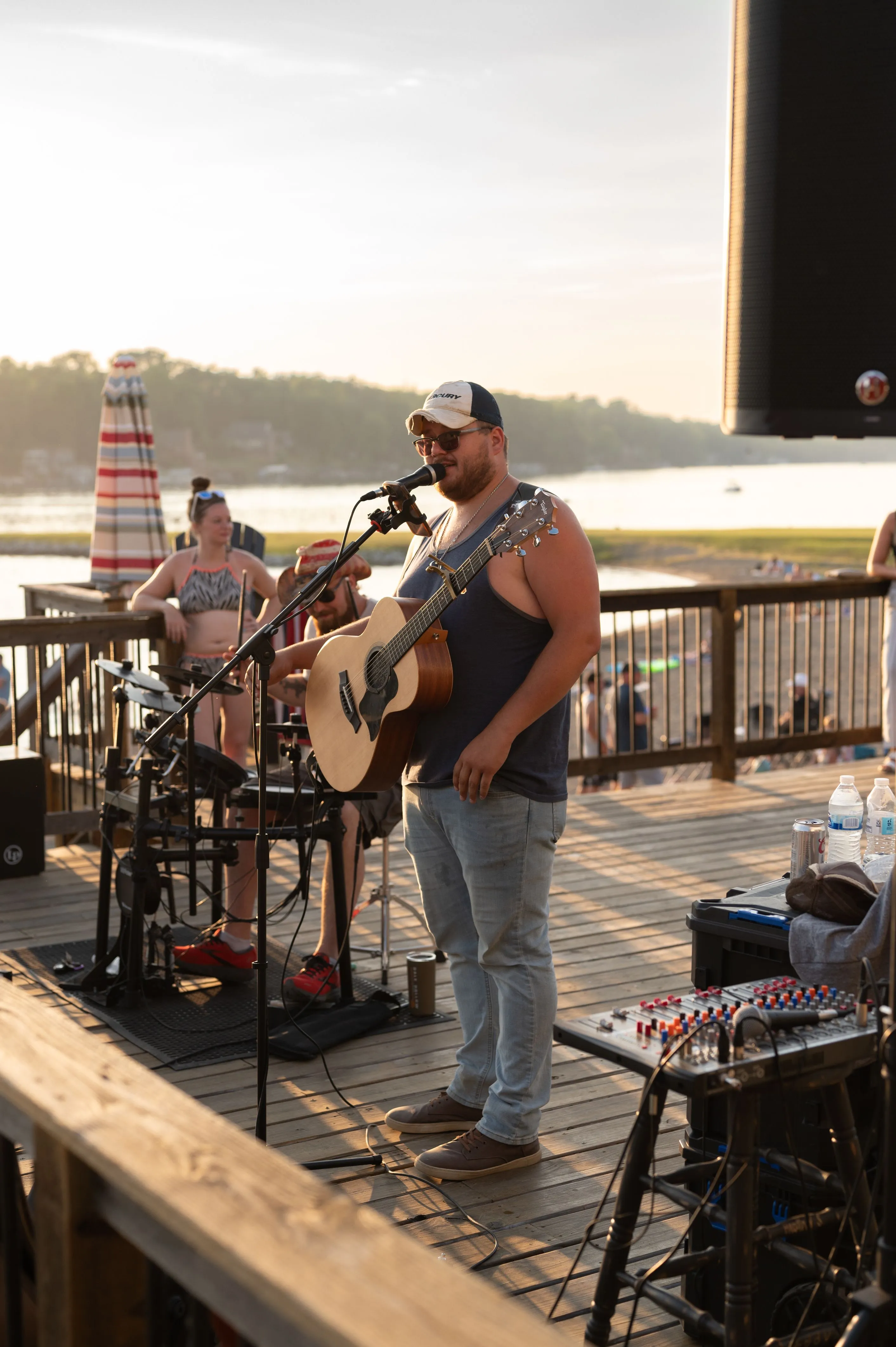 Man performing with a guitar on an outdoor deck overlooking a body of water with a lighthouse in the background.