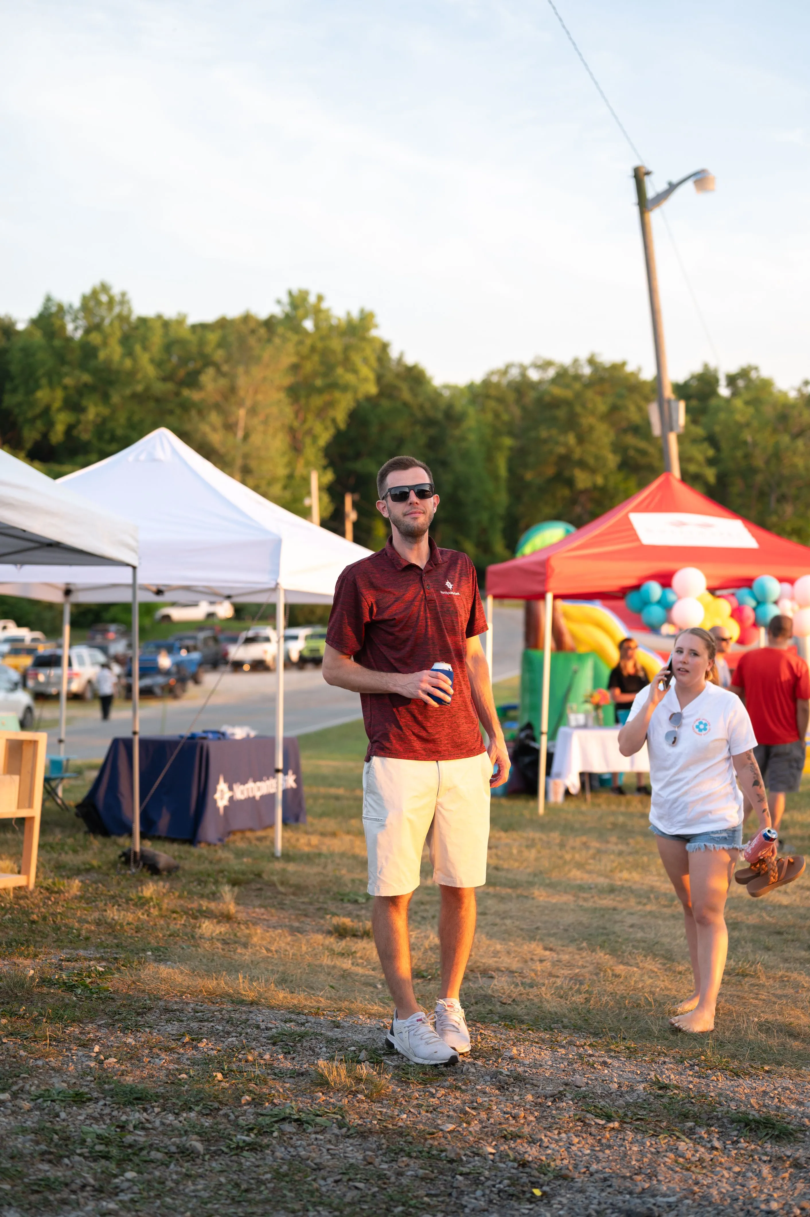 Man in sunglasses and a hat walking at an outdoor event with tents and people in the background.