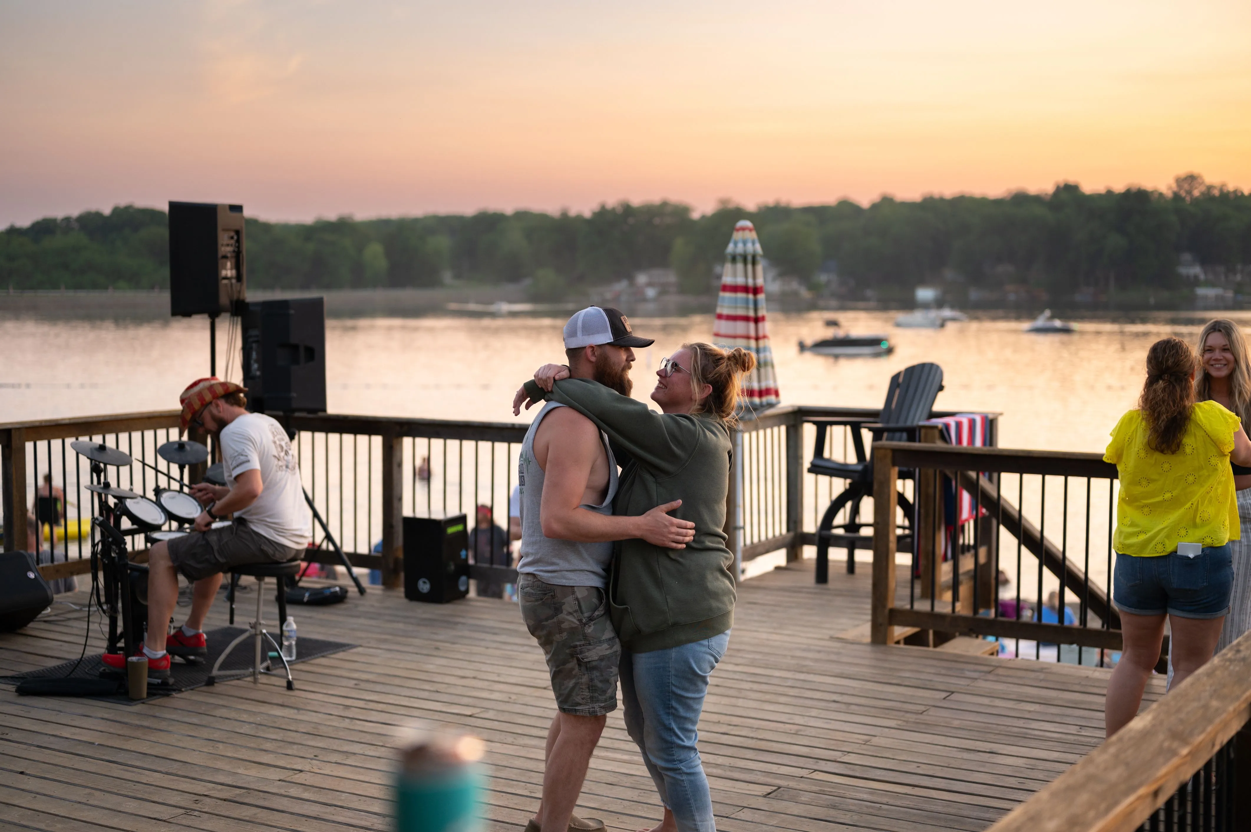 People enjoying a lakeside sunset, with a couple dancing on a deck and others relaxing near a speaker and overlooking the water.