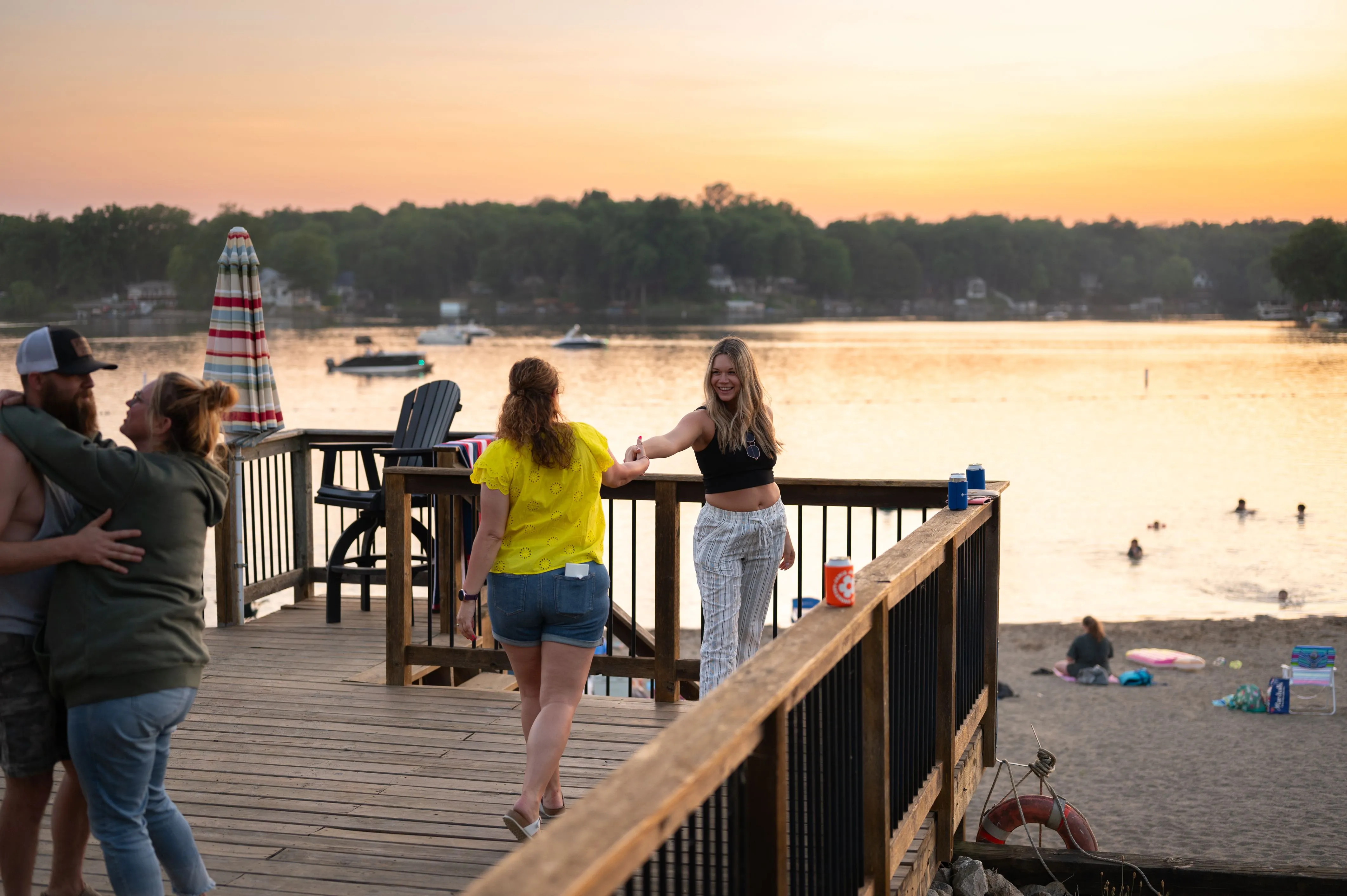 People enjoying a sunset at a lakeside dock, with some individuals standing, walking, and embracing.