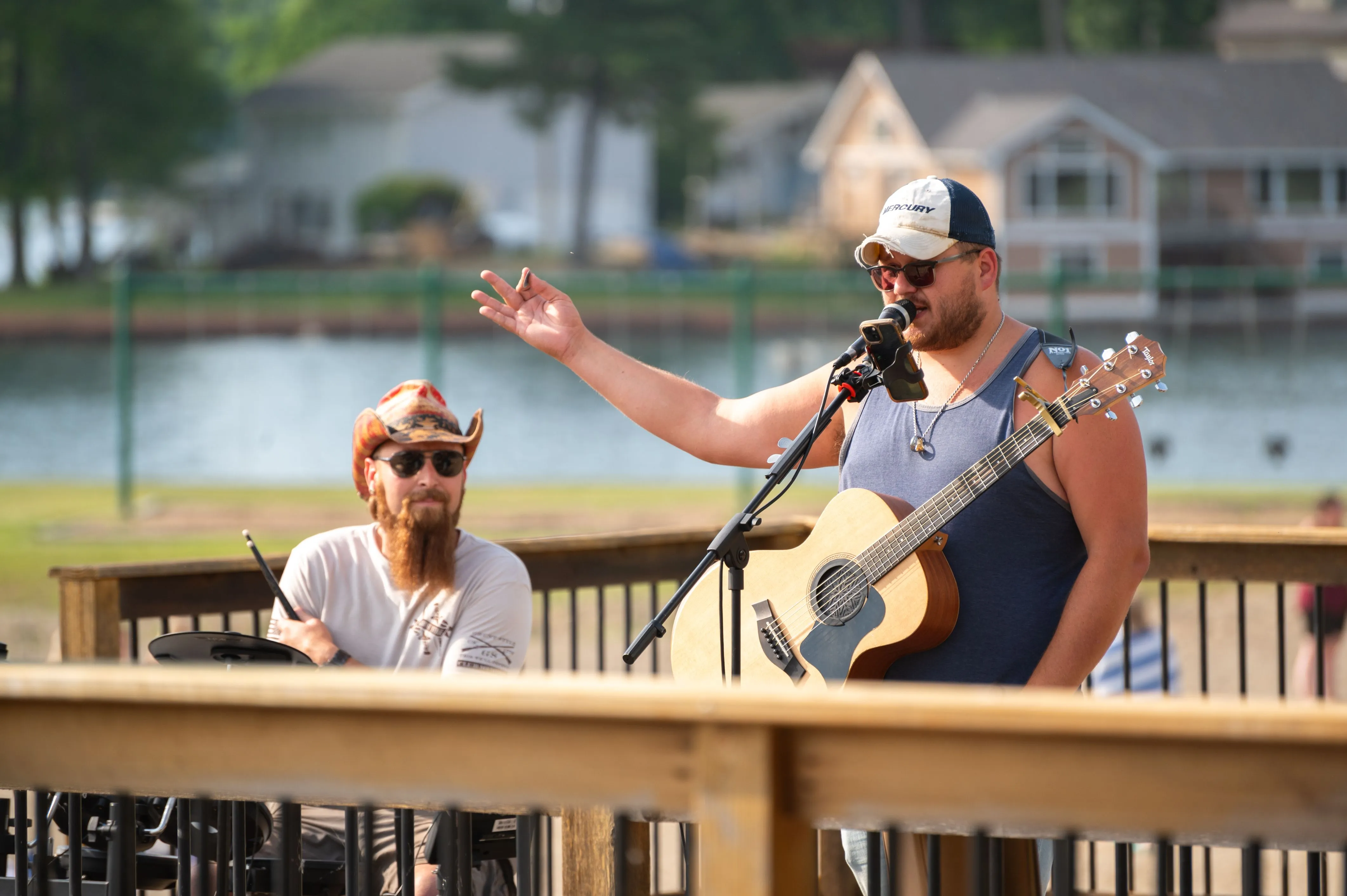 Two musicians performing on a wooden deck by a lake, one playing guitar and the other singing with a microphone.