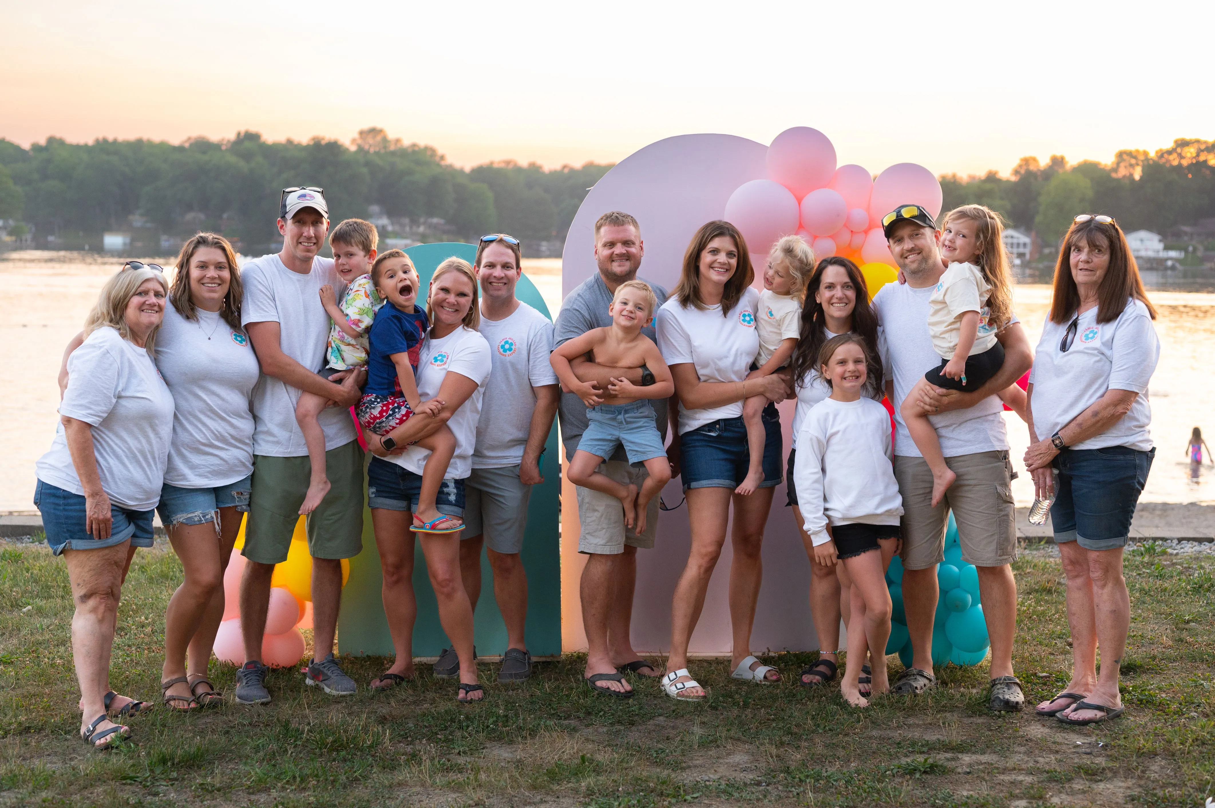 Group of people posing for a family photo outdoors near a lake at dusk, with some holding balloons.
