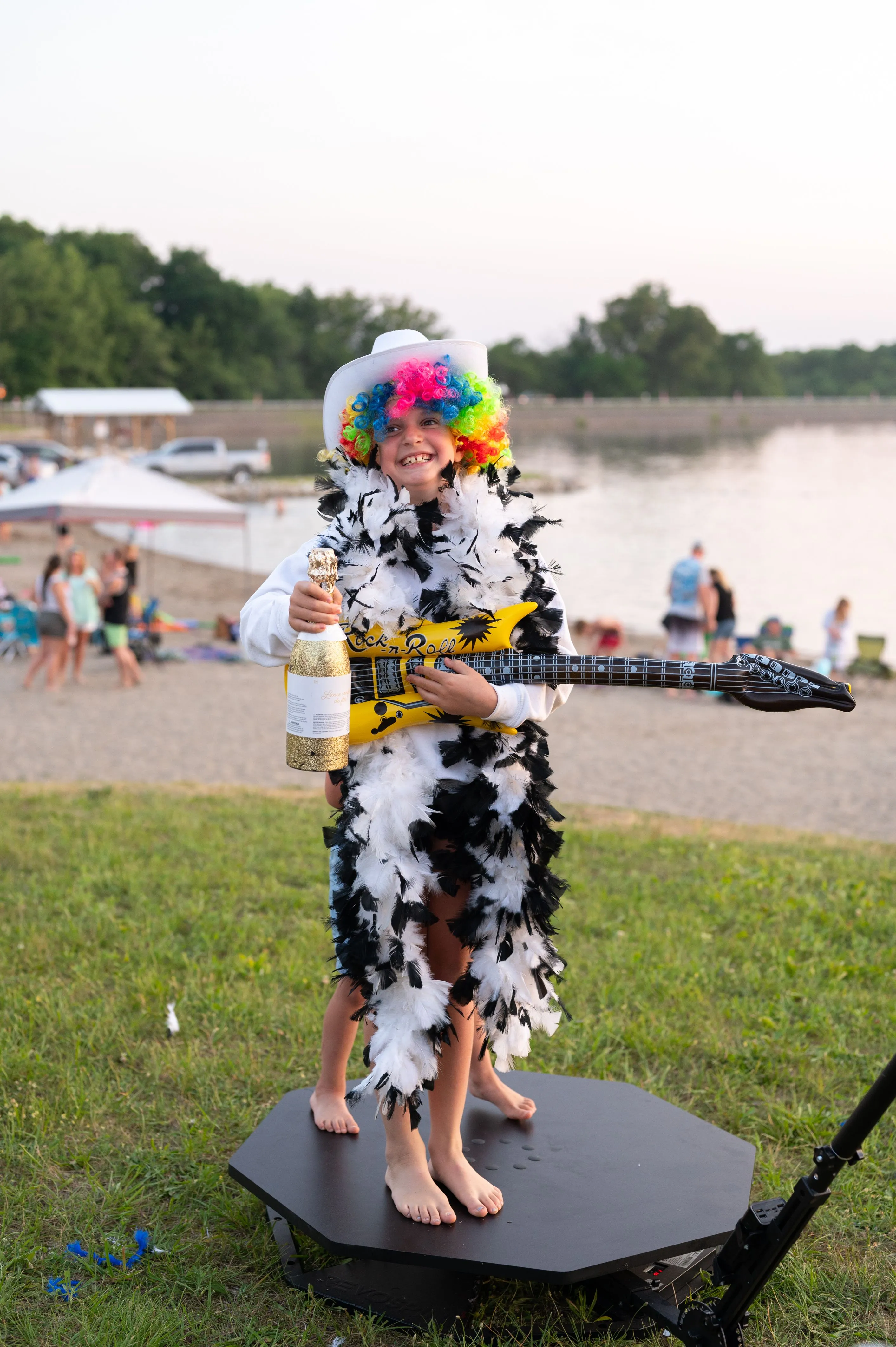 Person in a colorful costume with a hat and feathers, standing on an outdoor stage near a lake with people in the background.