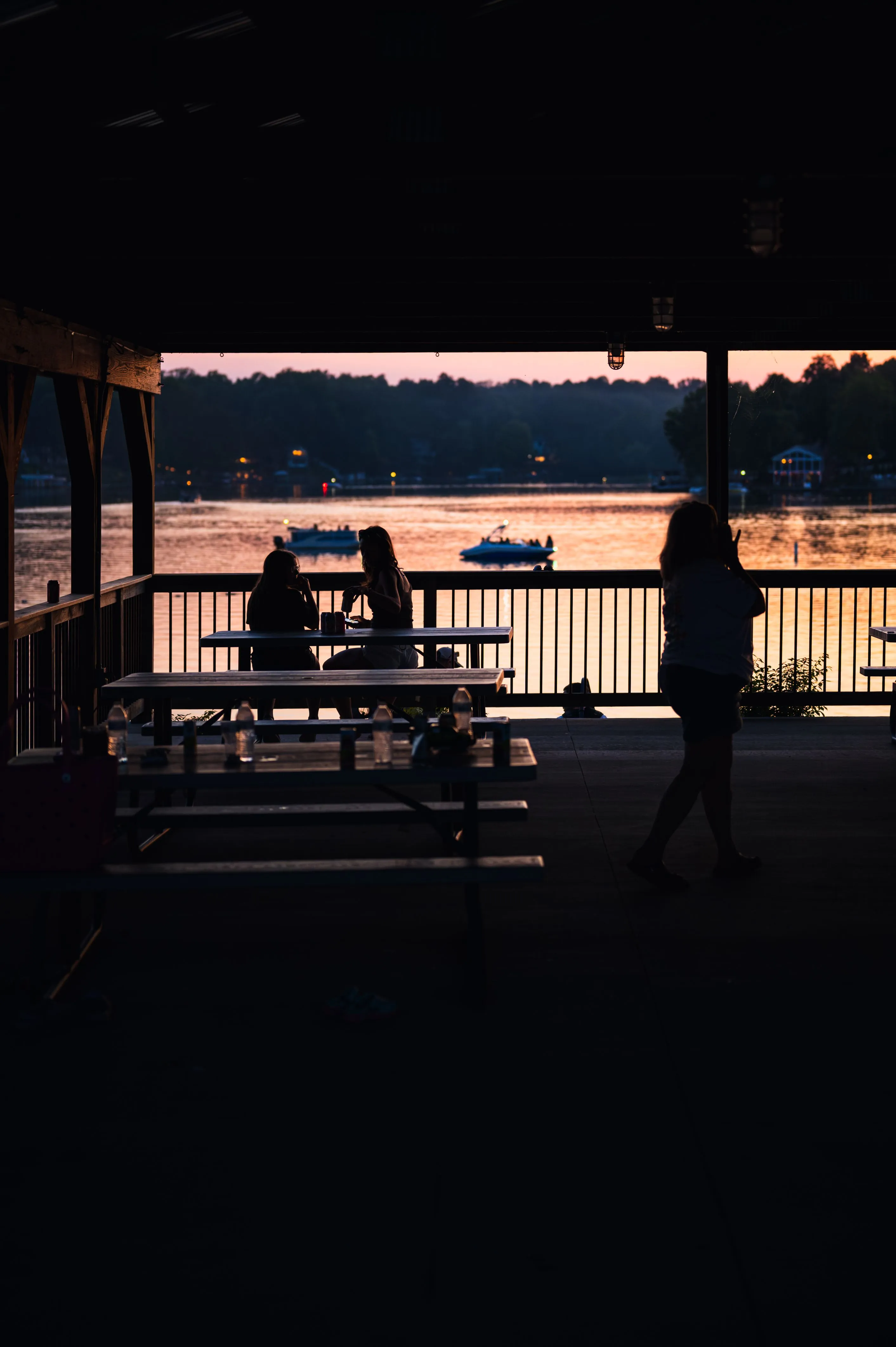 Silhouette of a person standing by a railing under a shelter with a distant sunset over a calm body of water.