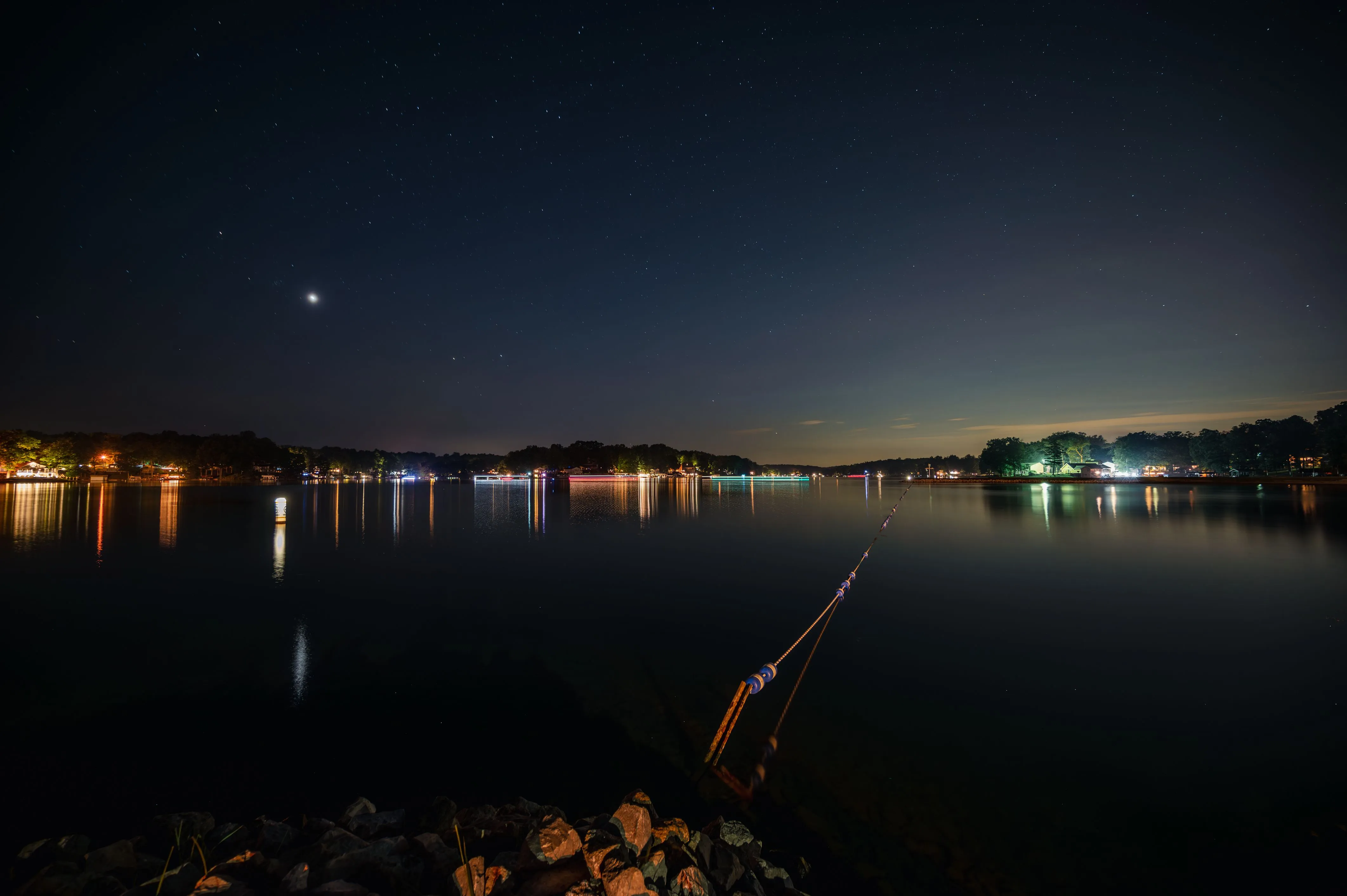 Nighttime view of a calm river reflecting lights from the opposite shore under a starry sky.