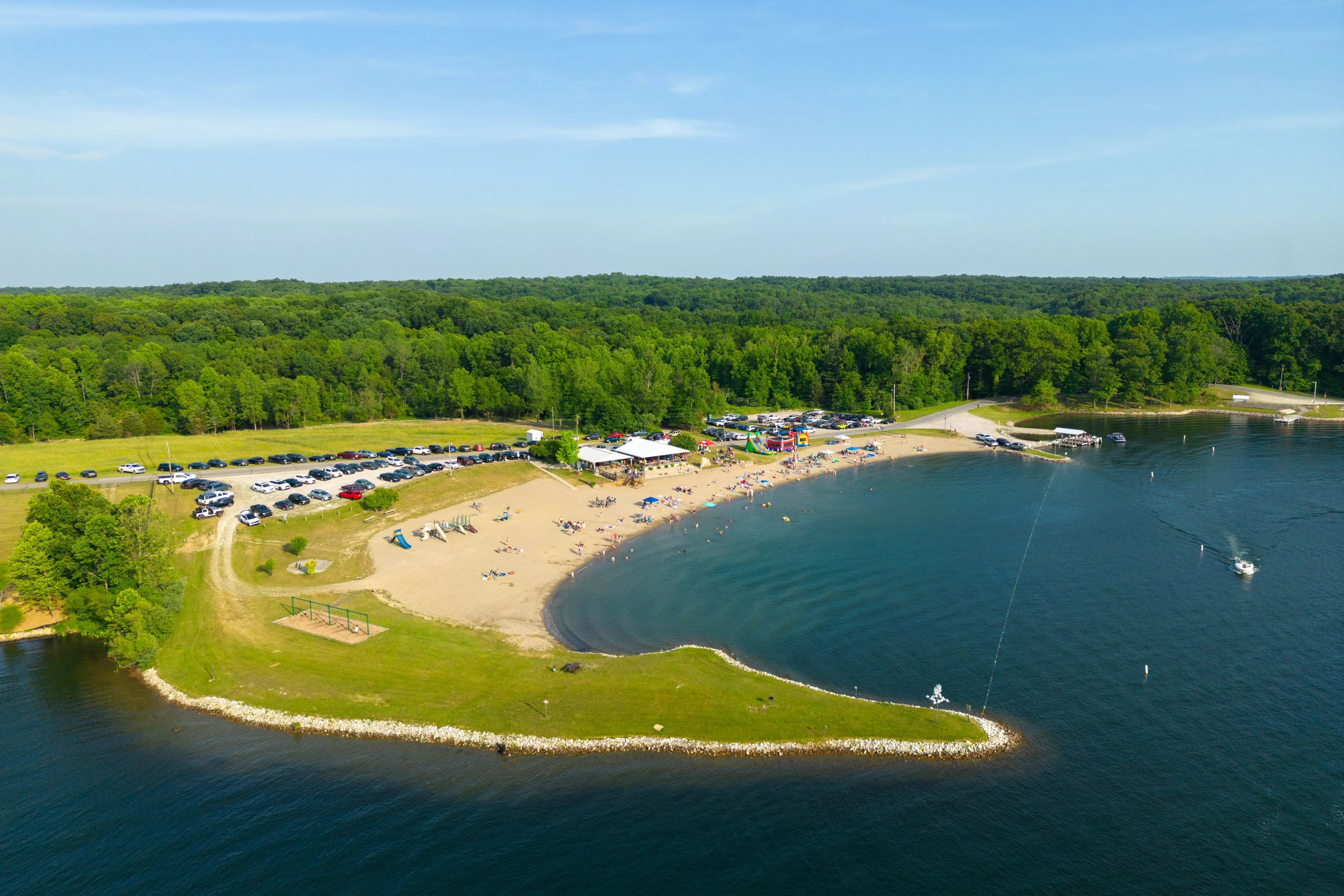 Aerial view of a beach park with a parking lot, trees, a sandy shoreline, and people enjoying water activities on a sunny day.
