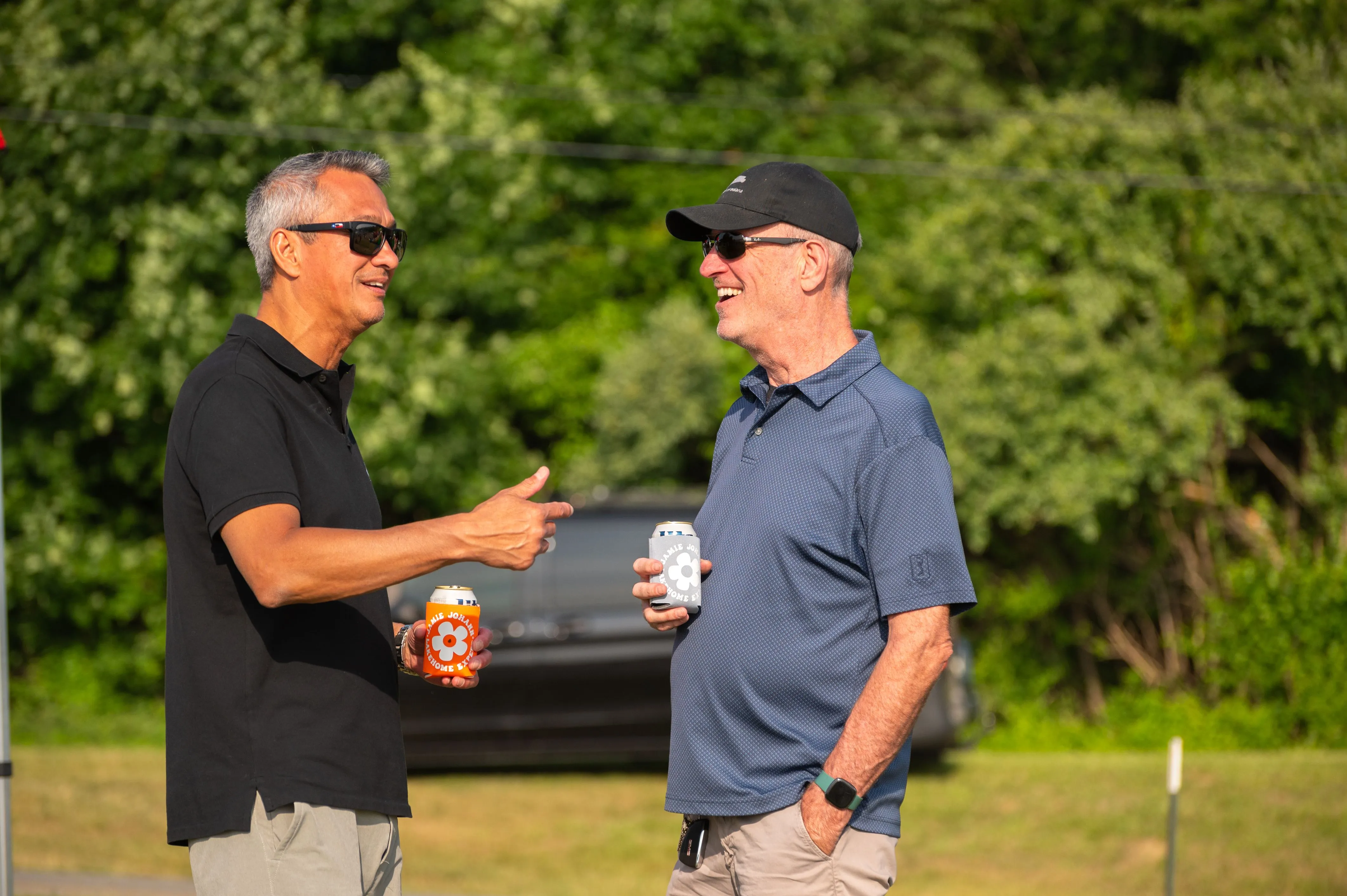 Two men talking outdoors with one pointing and both holding drinks, with trees in the background.
