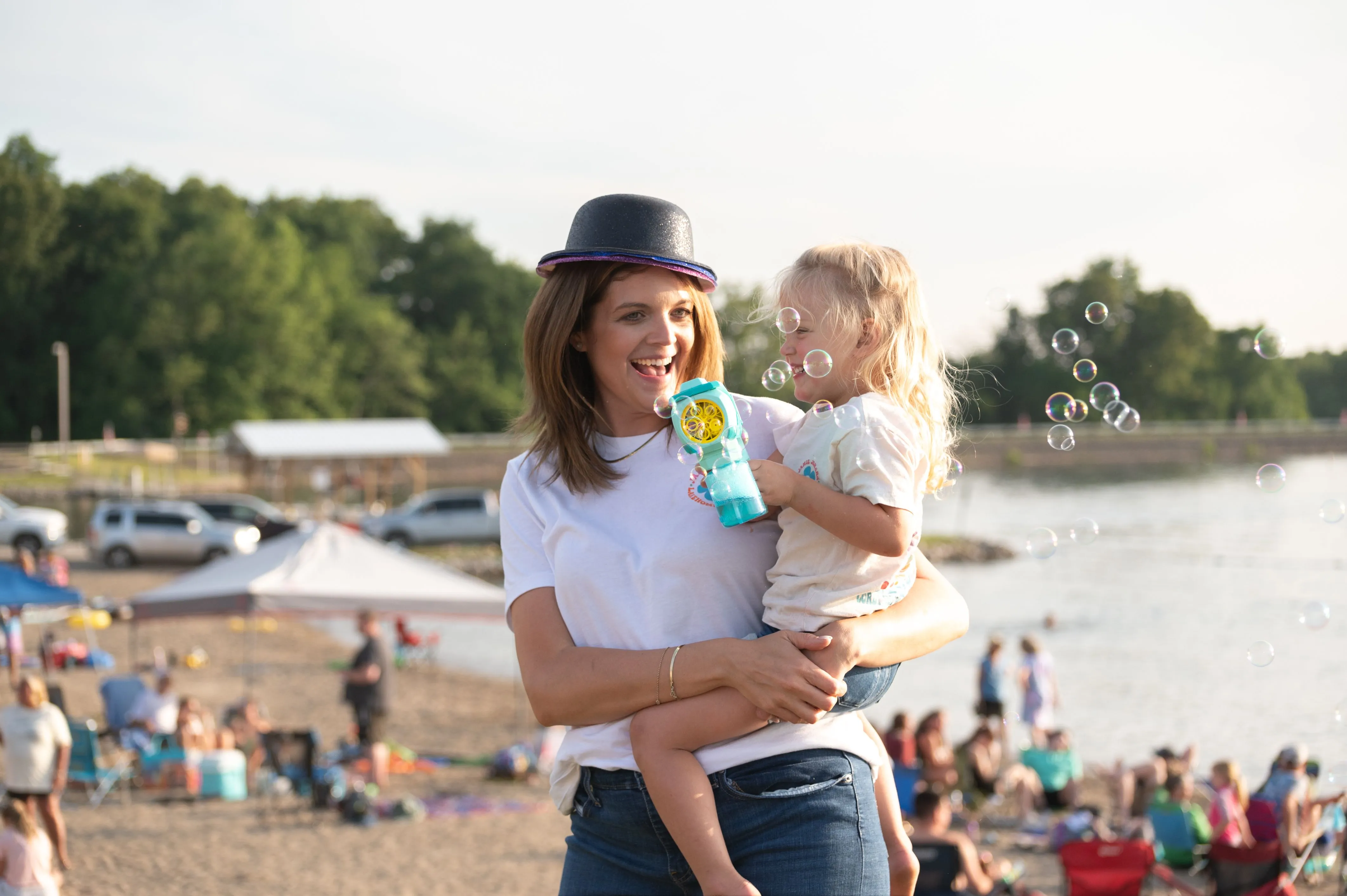 Woman in a hat holding a child blowing bubbles at a crowded beach.