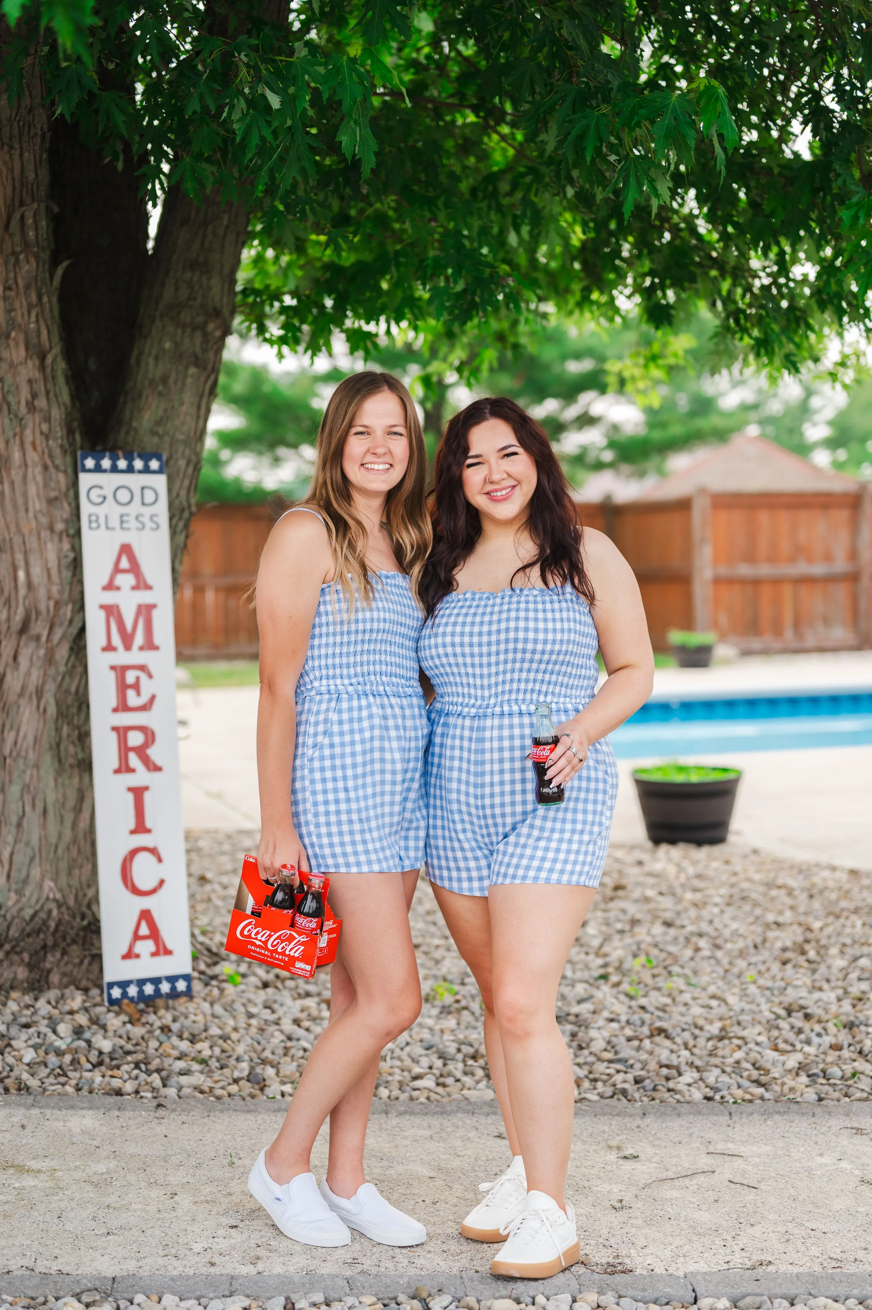 Two smiling women wearing matching blue and white checkered outfits standing by a tree with a "God Bless America" sign, one holding a Coca-Cola bottle, and the other holding a six-pack of Coca-Cola.