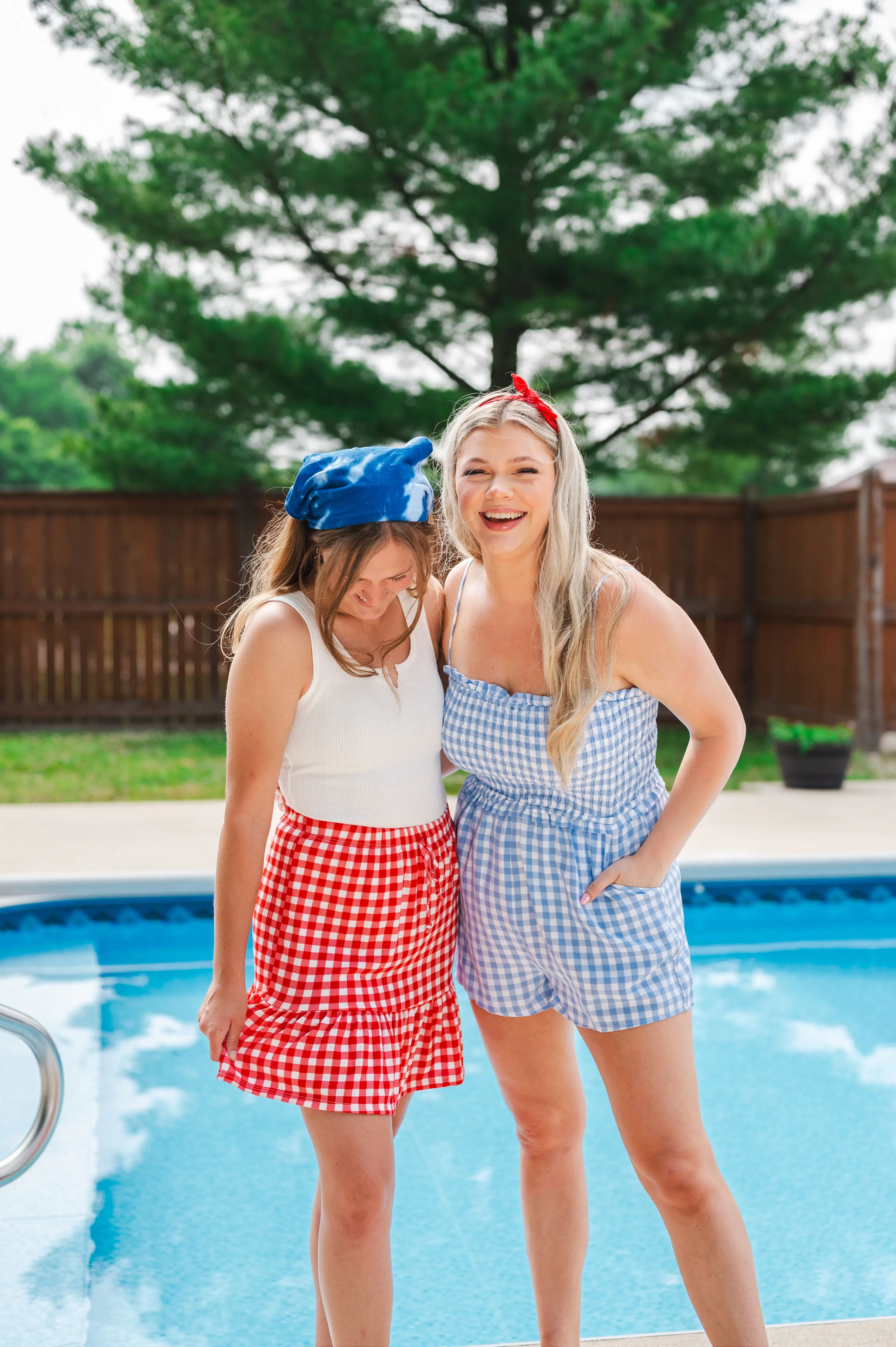 Two smiling young women standing by a pool, one wearing a red checkered skirt and the other in a blue checkered romper, with a plush dolphin on the head of the first woman.