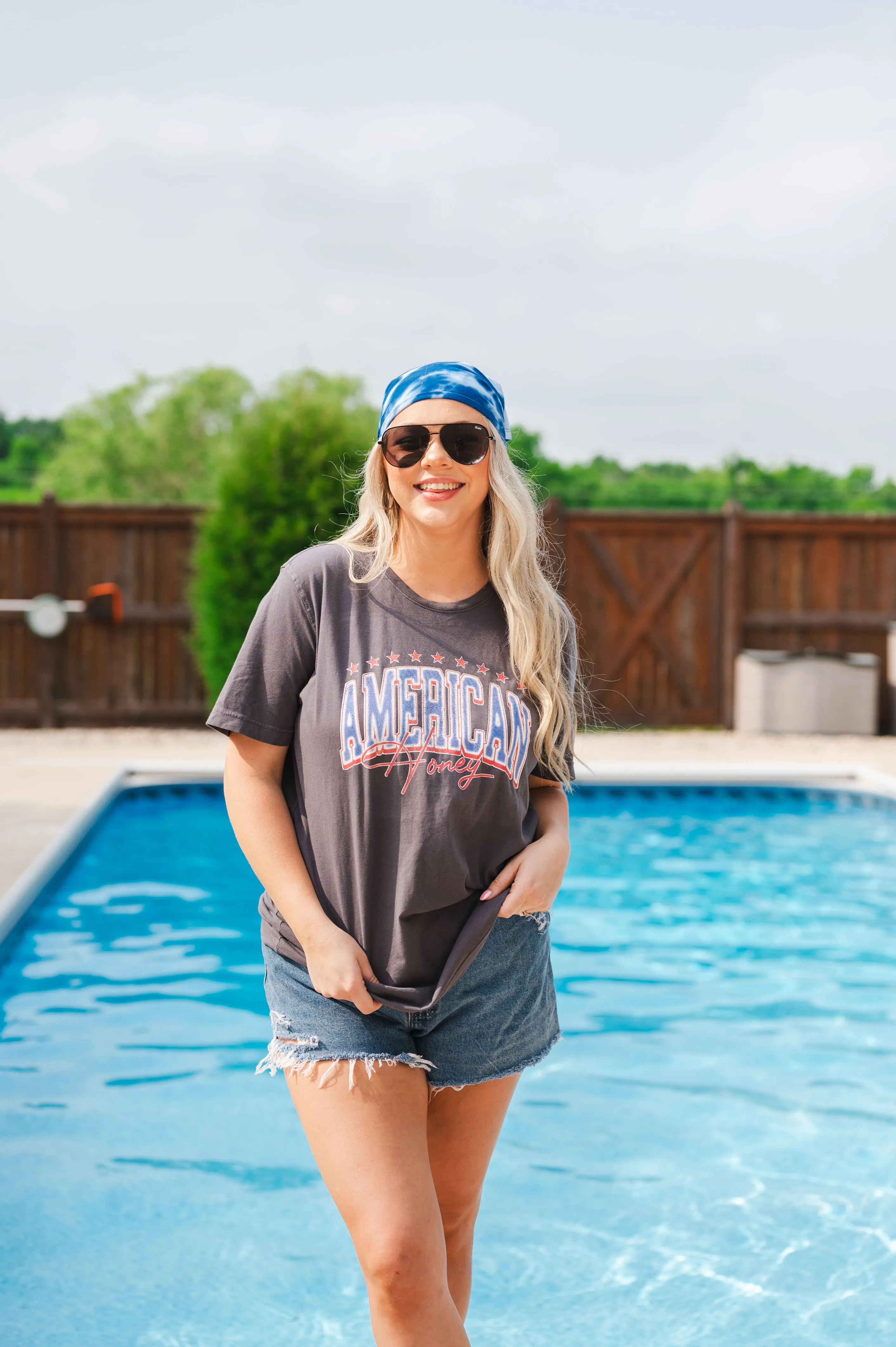Woman in a casual t-shirt and denim shorts standing by a swimming pool, wearing sunglasses and a blue bandana on her head.