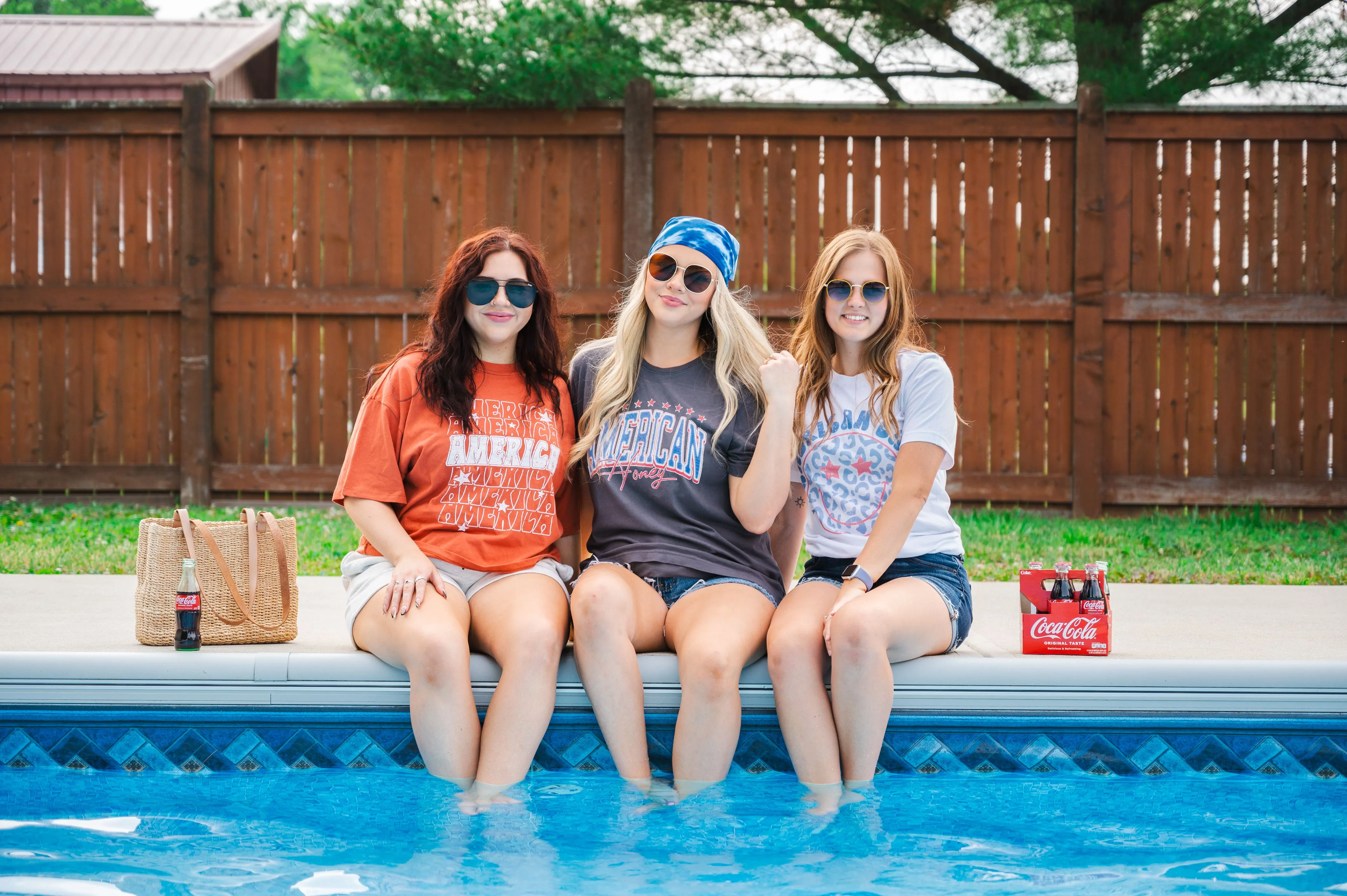 Three women sitting by a pool with their feet dipped in the water, smiling, wearing sunglasses, and patriotic-themed T-shirts. A woven bag and a six-pack of Coca-Cola are placed on the poolside. Wooden fence and greenery are visible in the background.