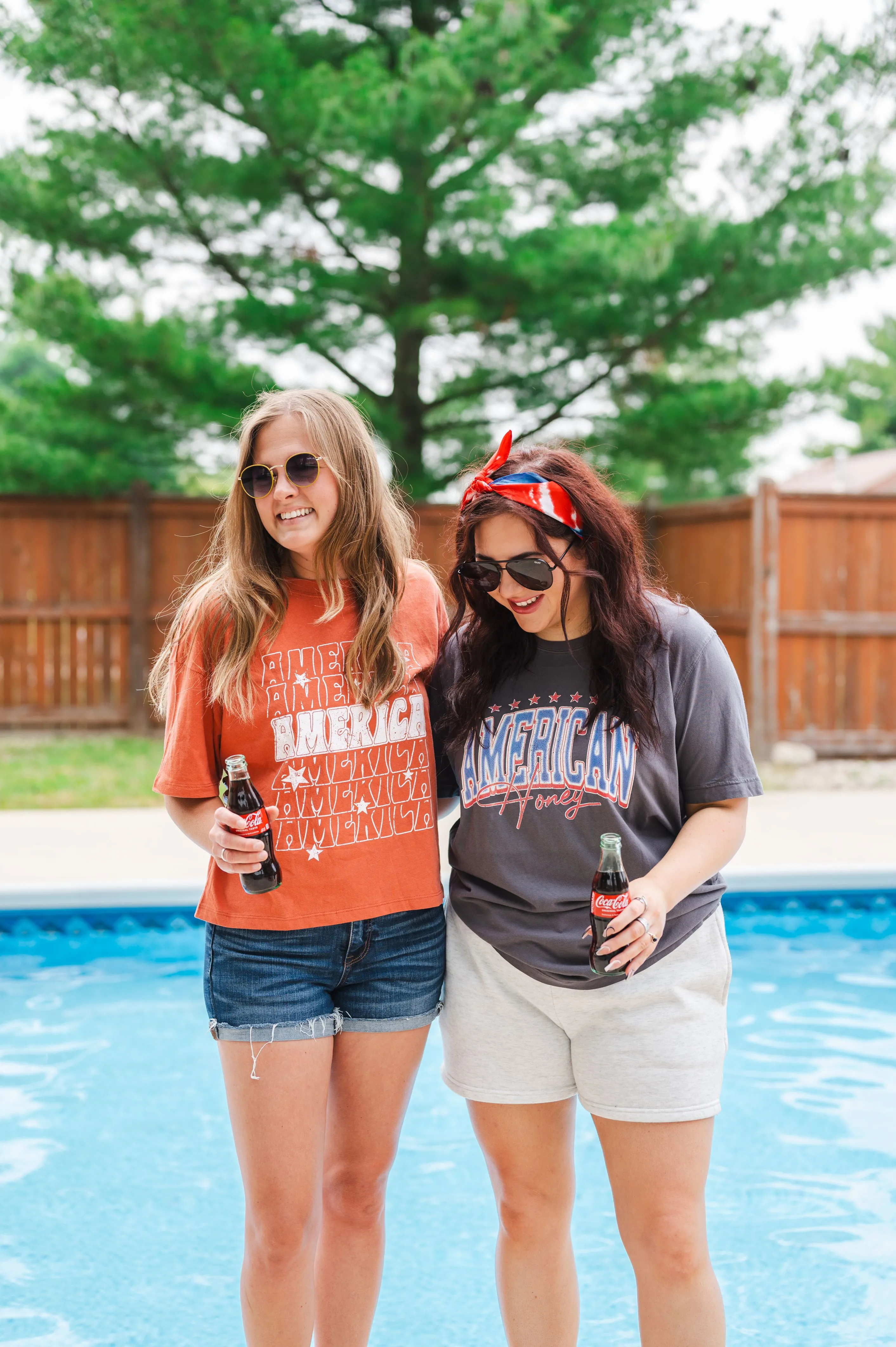 Two women smiling and holding soda bottles by a poolside, dressed in casual summer clothes with patriotic themes.
