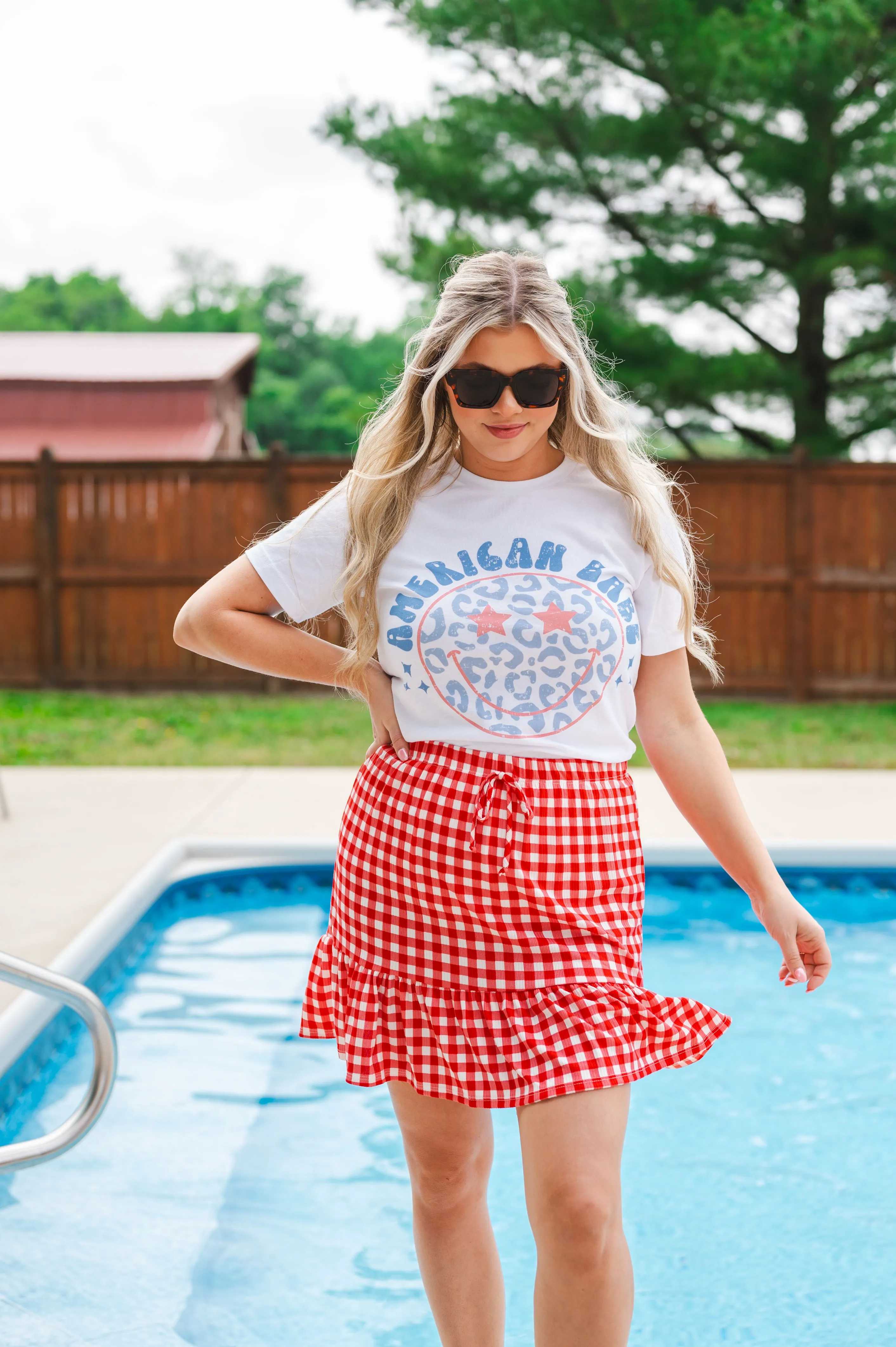 Woman in sunglasses posing by the pool wearing a white t-shirt with 'American Babe' print and red checked skirt.