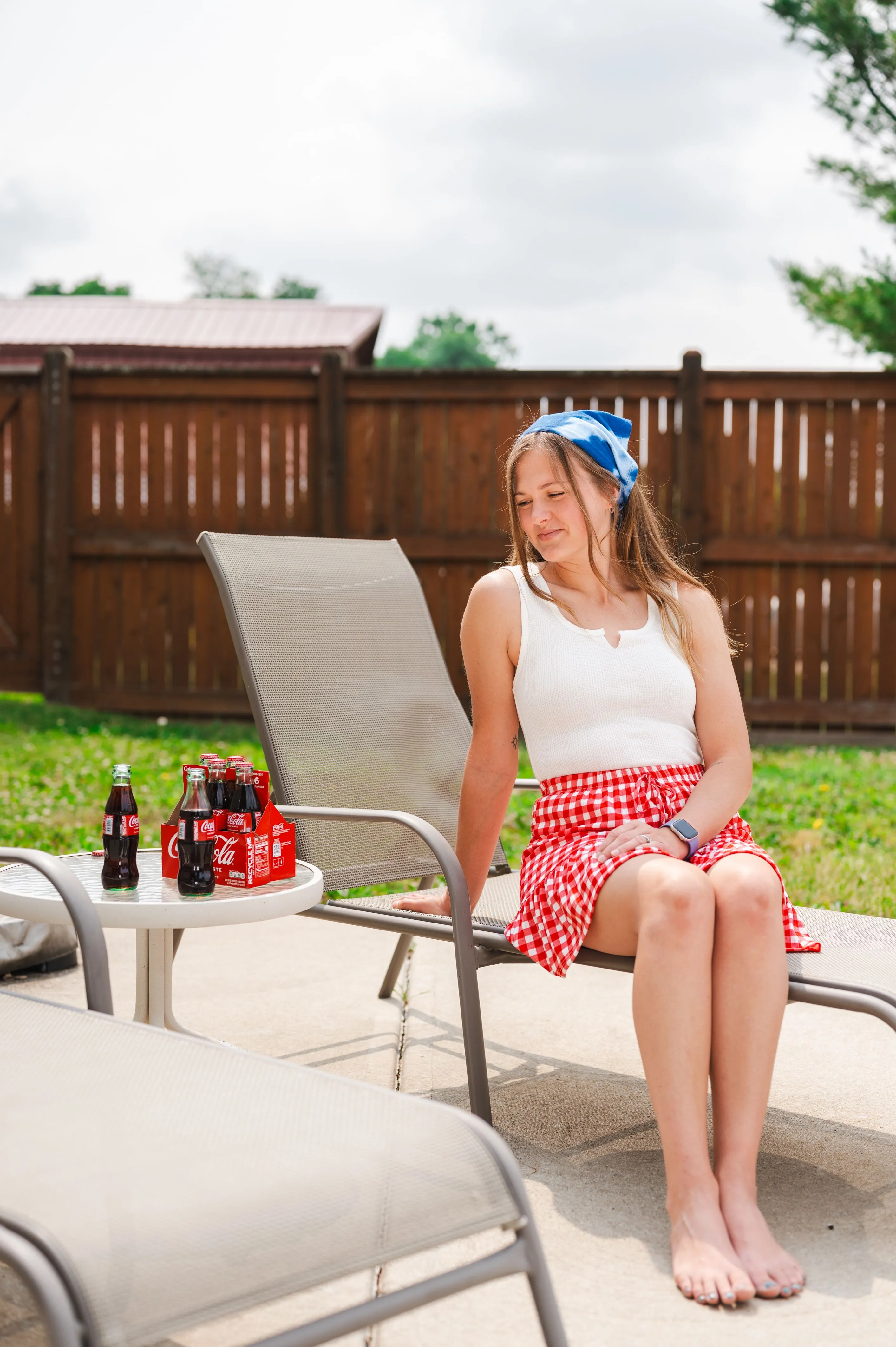 Woman in a white tank top and red checkered skirt sitting outside next to a table with bottles of Coca-Cola.