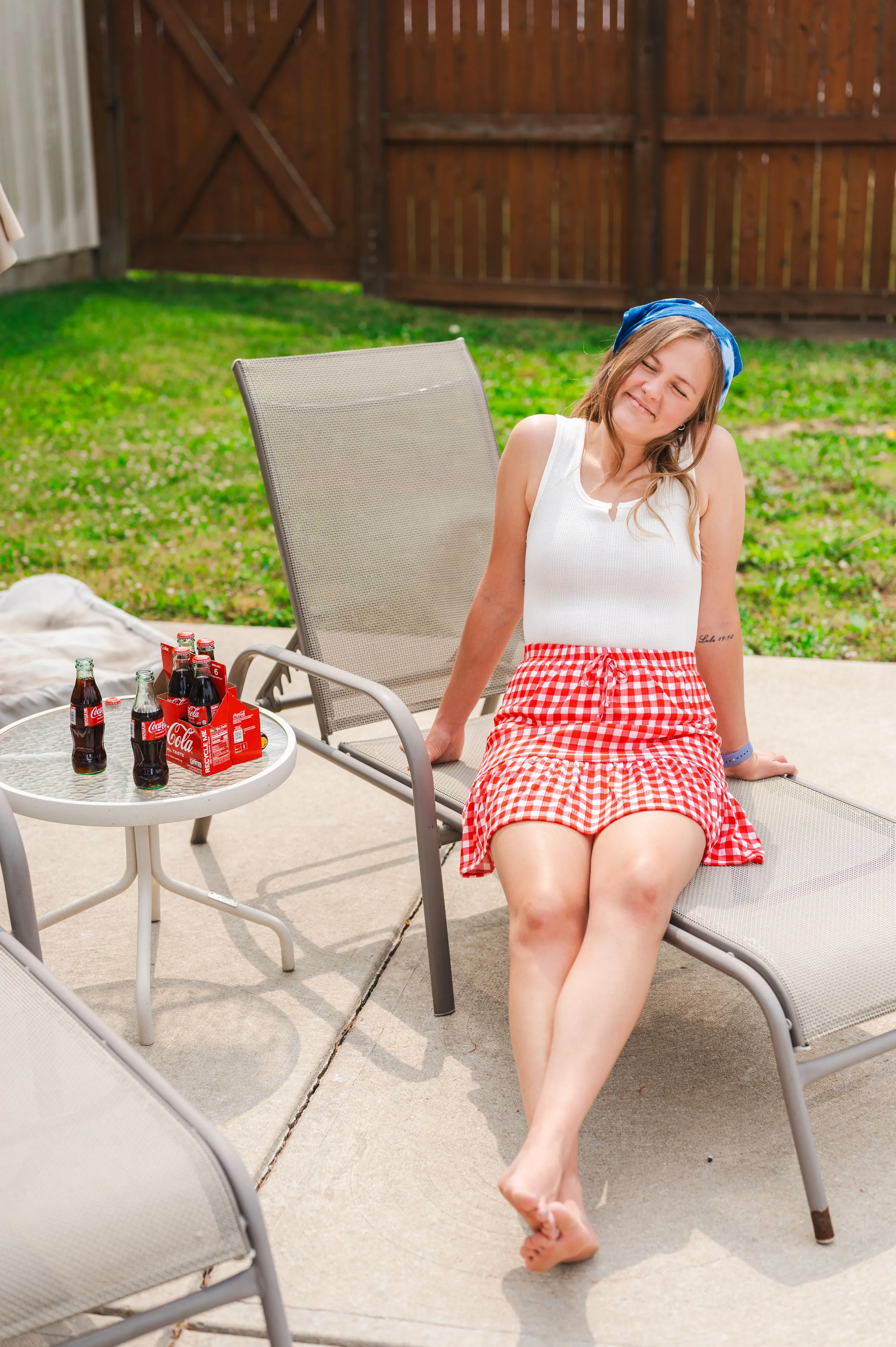 Woman smiling and relaxing on a patio chair with closed eyes, wearing a white tank top and red gingham skirt, with a table holding bottles of Coca-Cola nearby.