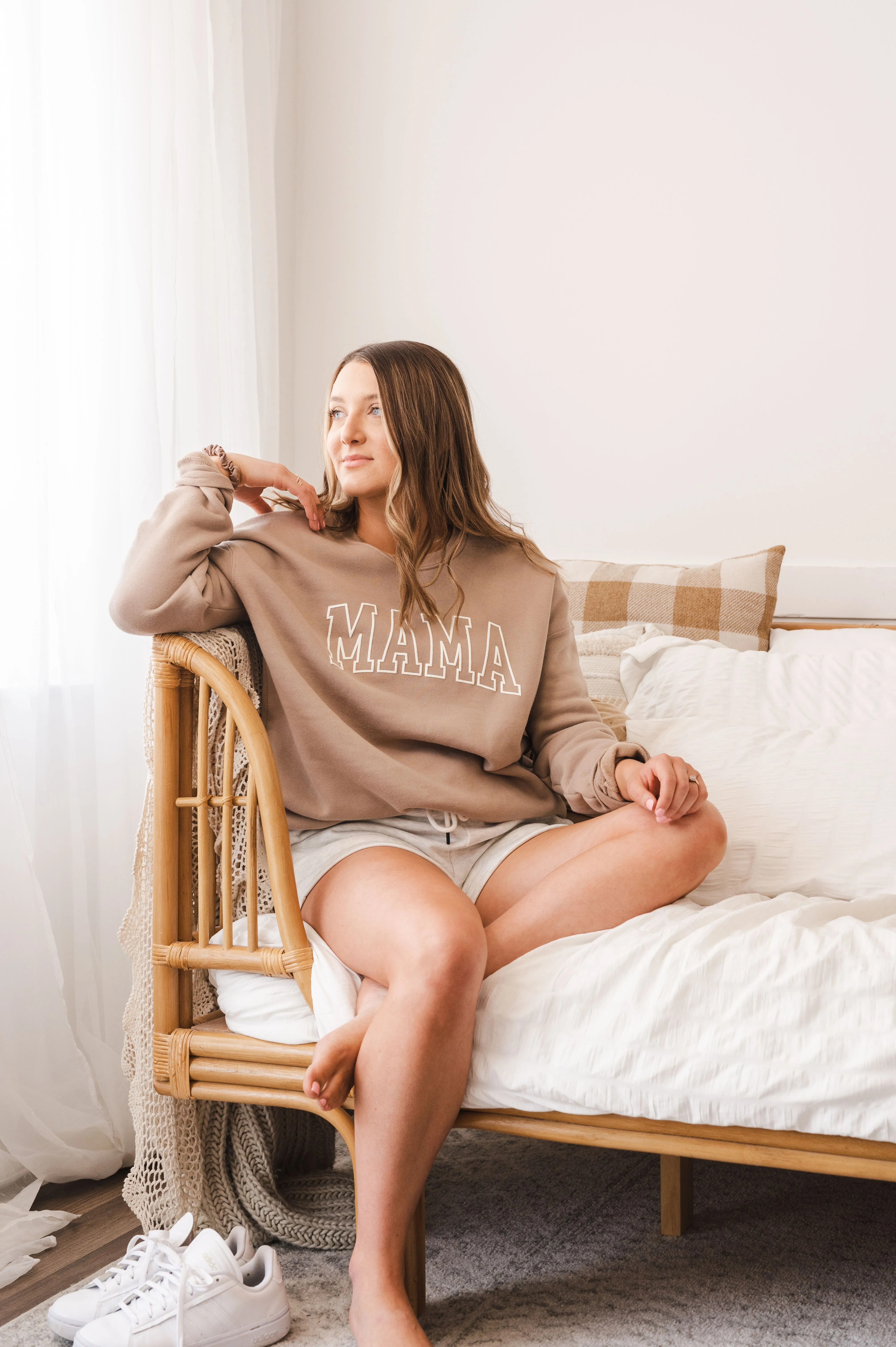 Woman sitting on a rattan chair in a cozy bedroom, wearing a "MAMA" sweatshirt and shorts, with white sneakers on the floor.