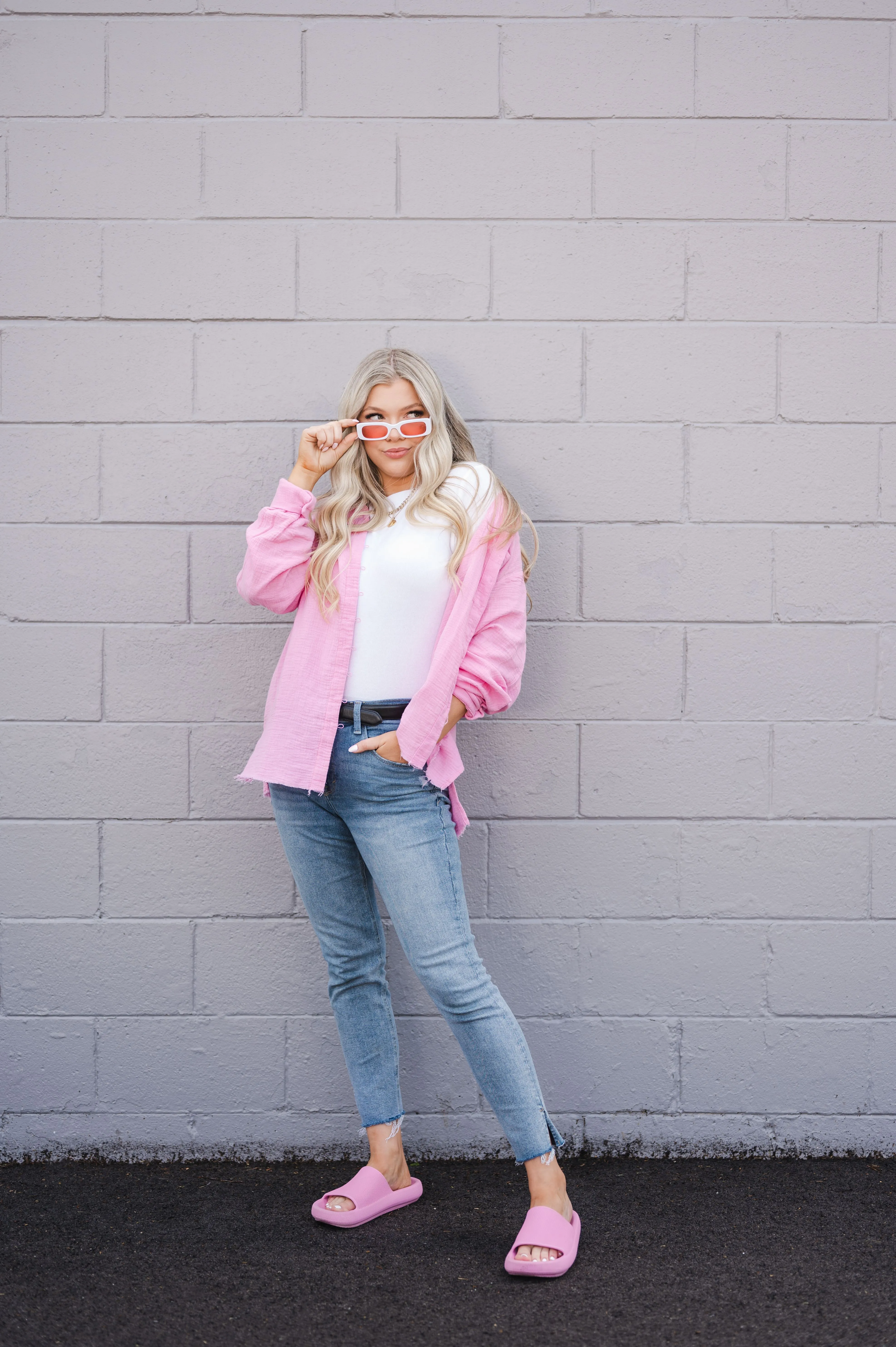 A woman in a pink jacket and blue jeans standing against a gray brick wall, playfully adjusting her white sunglasses.