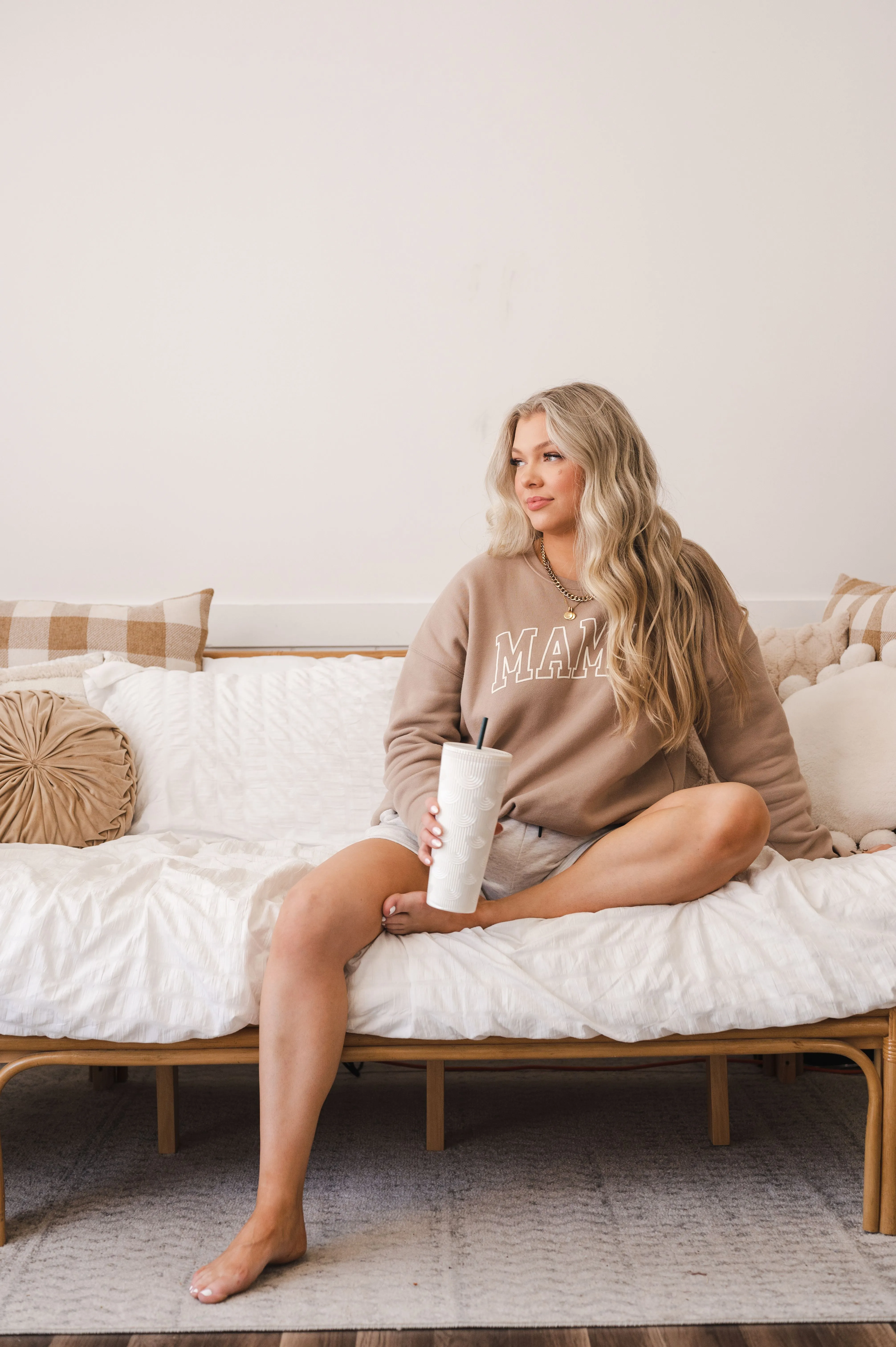 Woman sitting on a bed holding a tumbler, with a serene expression, in a cozy room environment.
