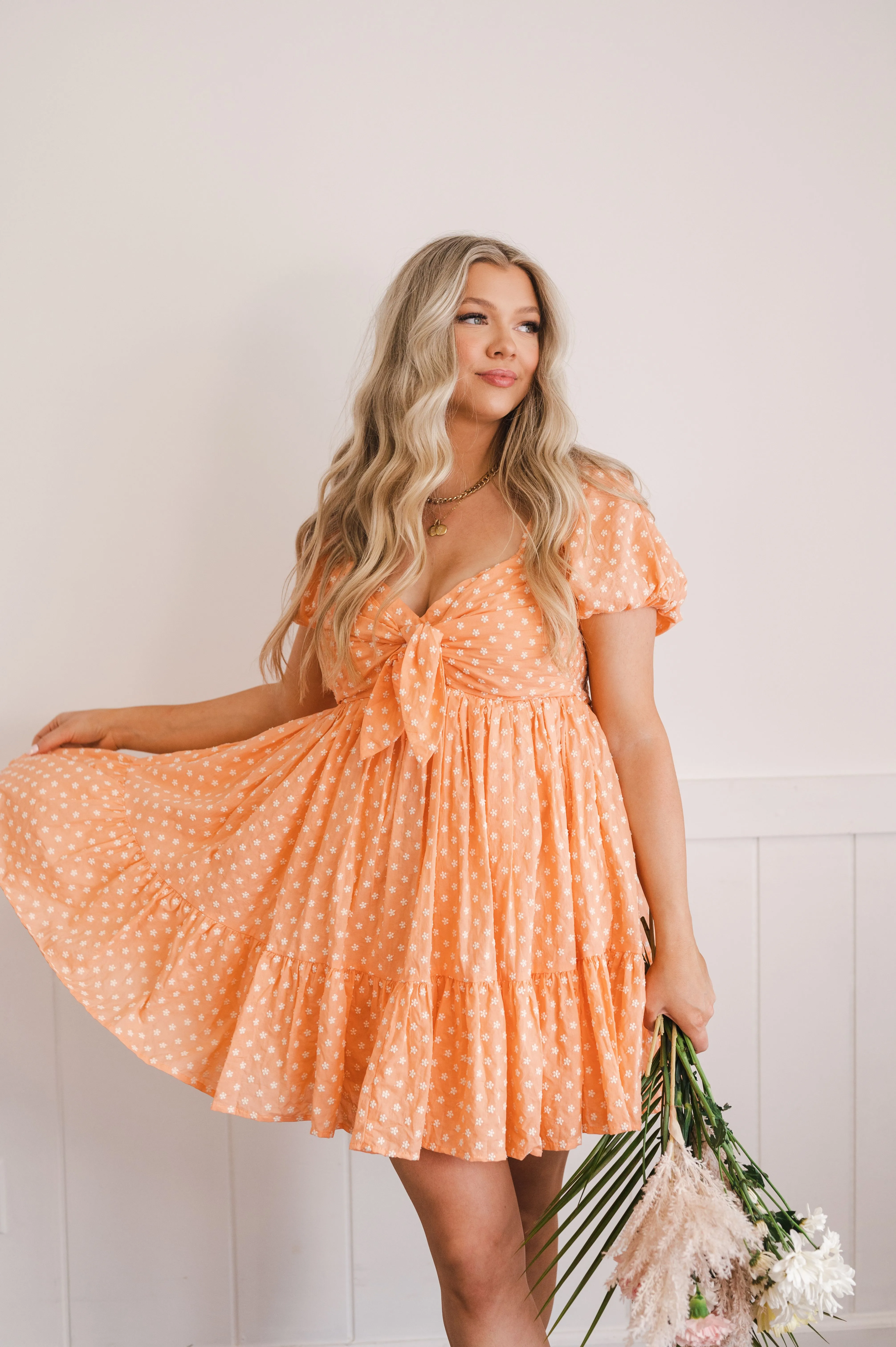 Woman in a peach polka dot dress holding flowers and posing with a twirl against a light background.