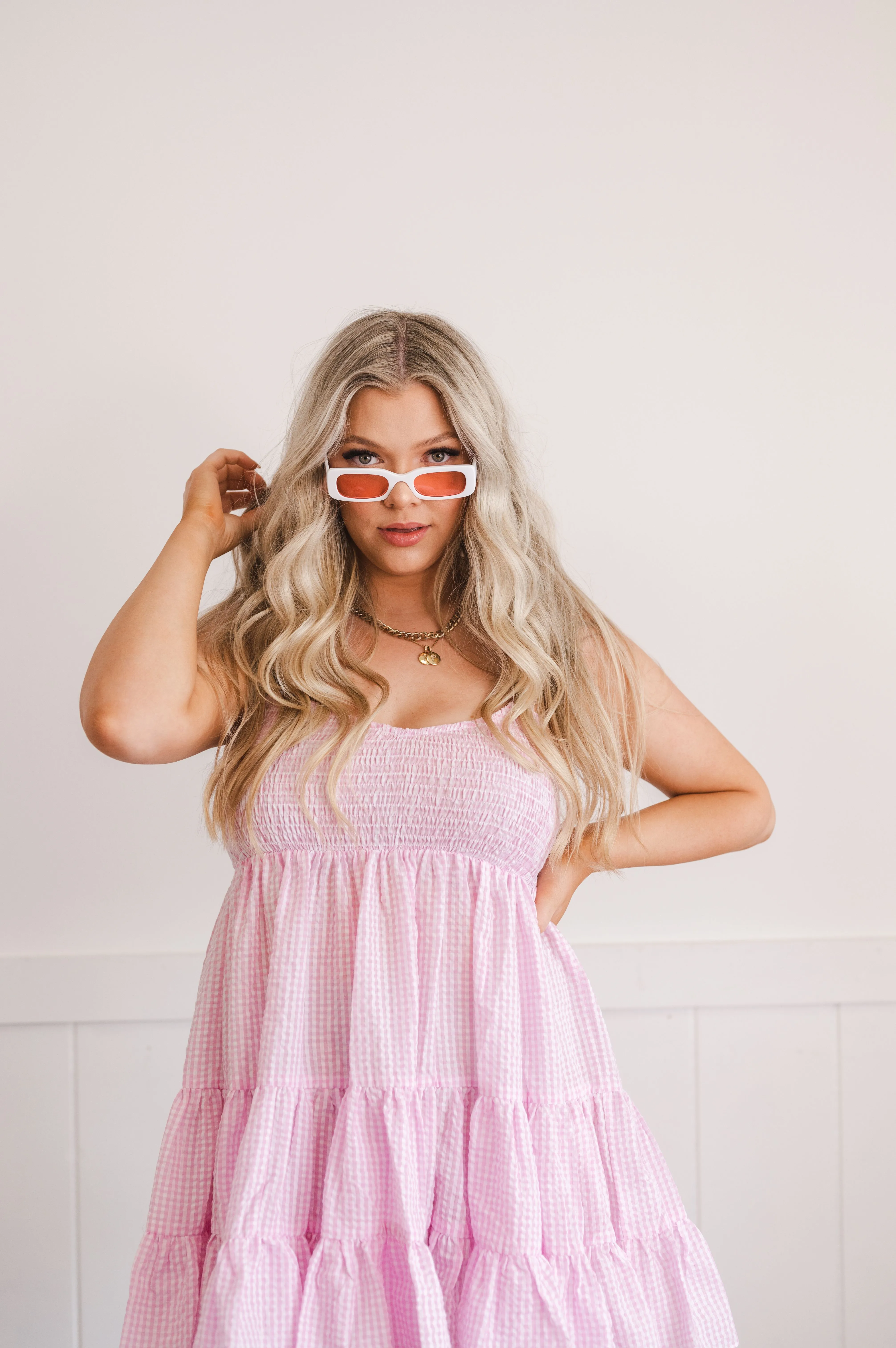 Woman in a pink gingham dress posing with white sunglasses against a light background.