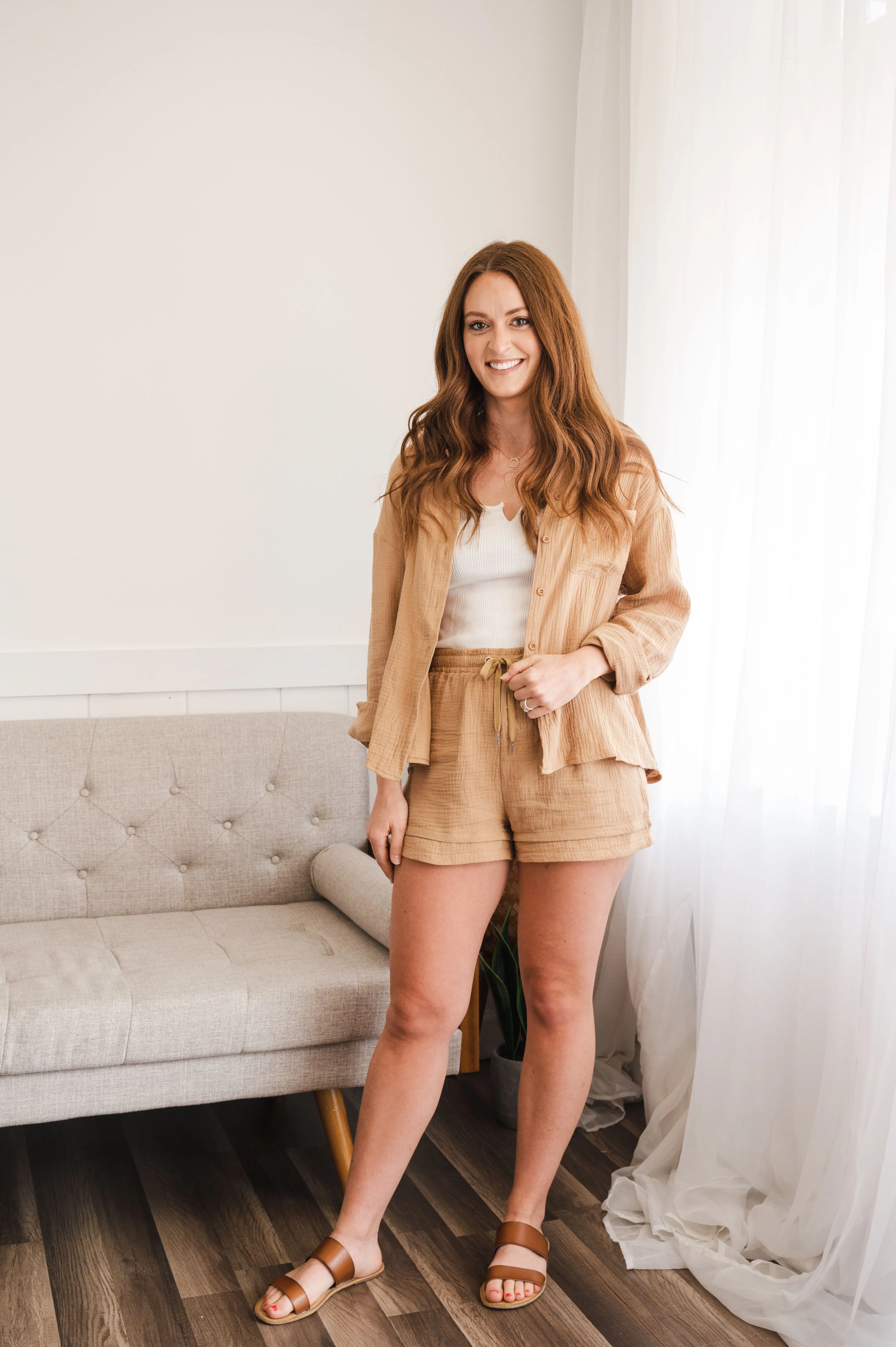 Young woman standing in a well-lit room wearing a casual tan-colored suit with shorts, white top, and sandals, smiling at the camera with a sheer curtain and a beige couch in the background.