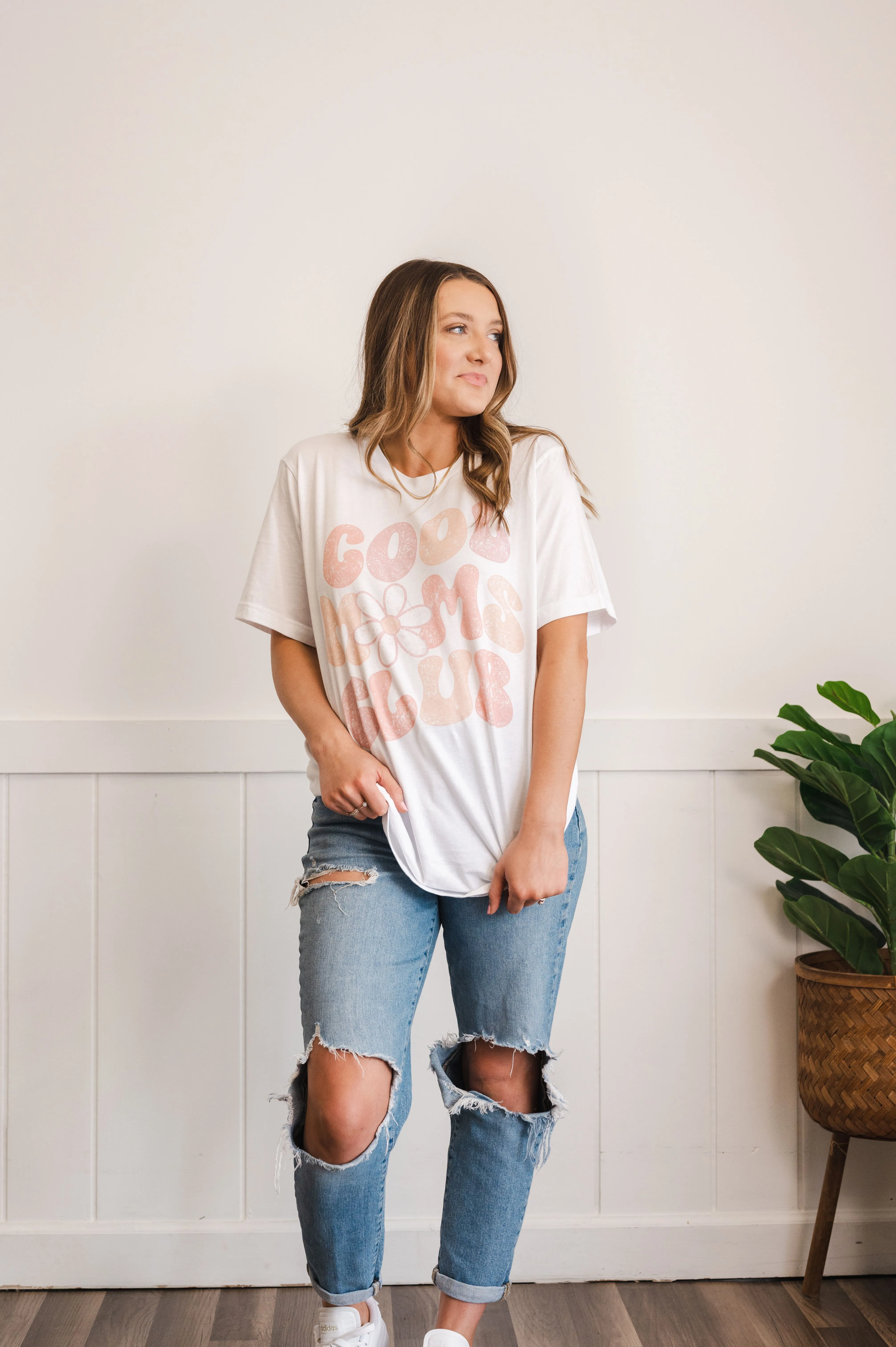 Woman in a white t-shirt with 'Good Vibes Club' print posing in ripped blue jeans, white sneakers, standing beside a potted plant against a white wall.