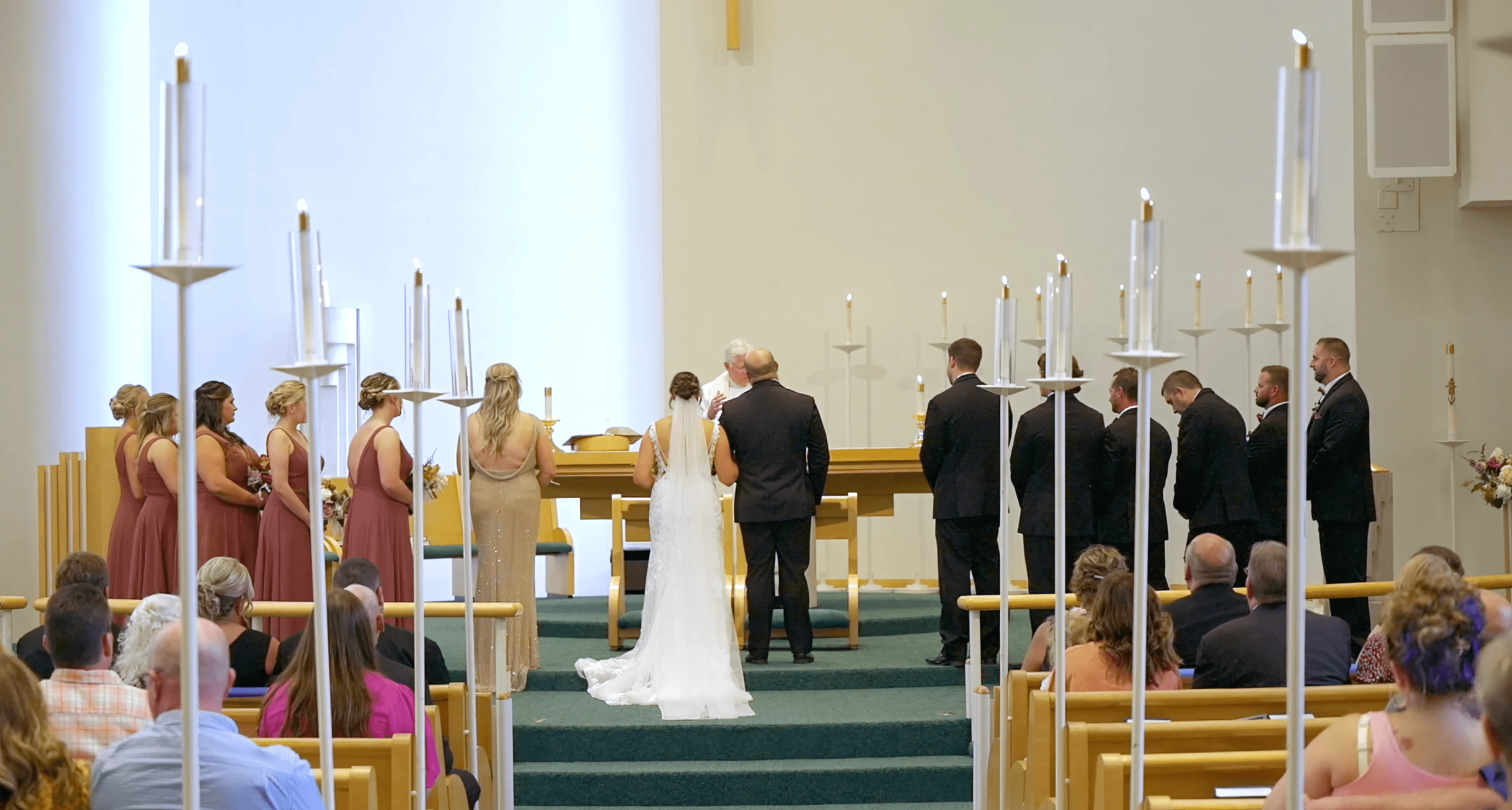 Wedding ceremony in a church with a bride and groom at the altar, bridesmaids in rose dresses, groomsmen in black suits, and guests seated in pews.