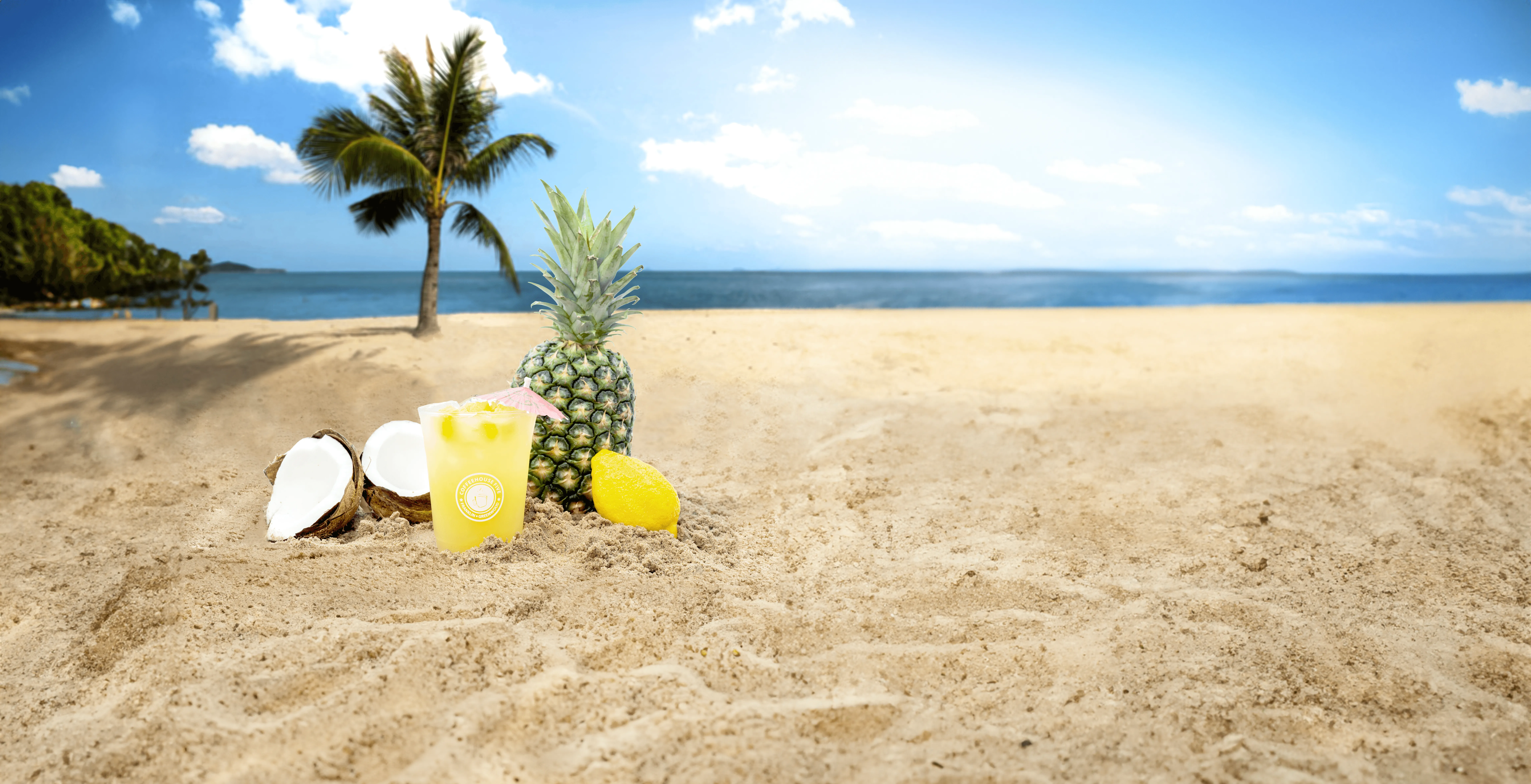 Tropical beach scene with a pineapple, coconut halves, a glass of lemonade, and a lemon slice on the sand, palm tree and blue sky in the background.