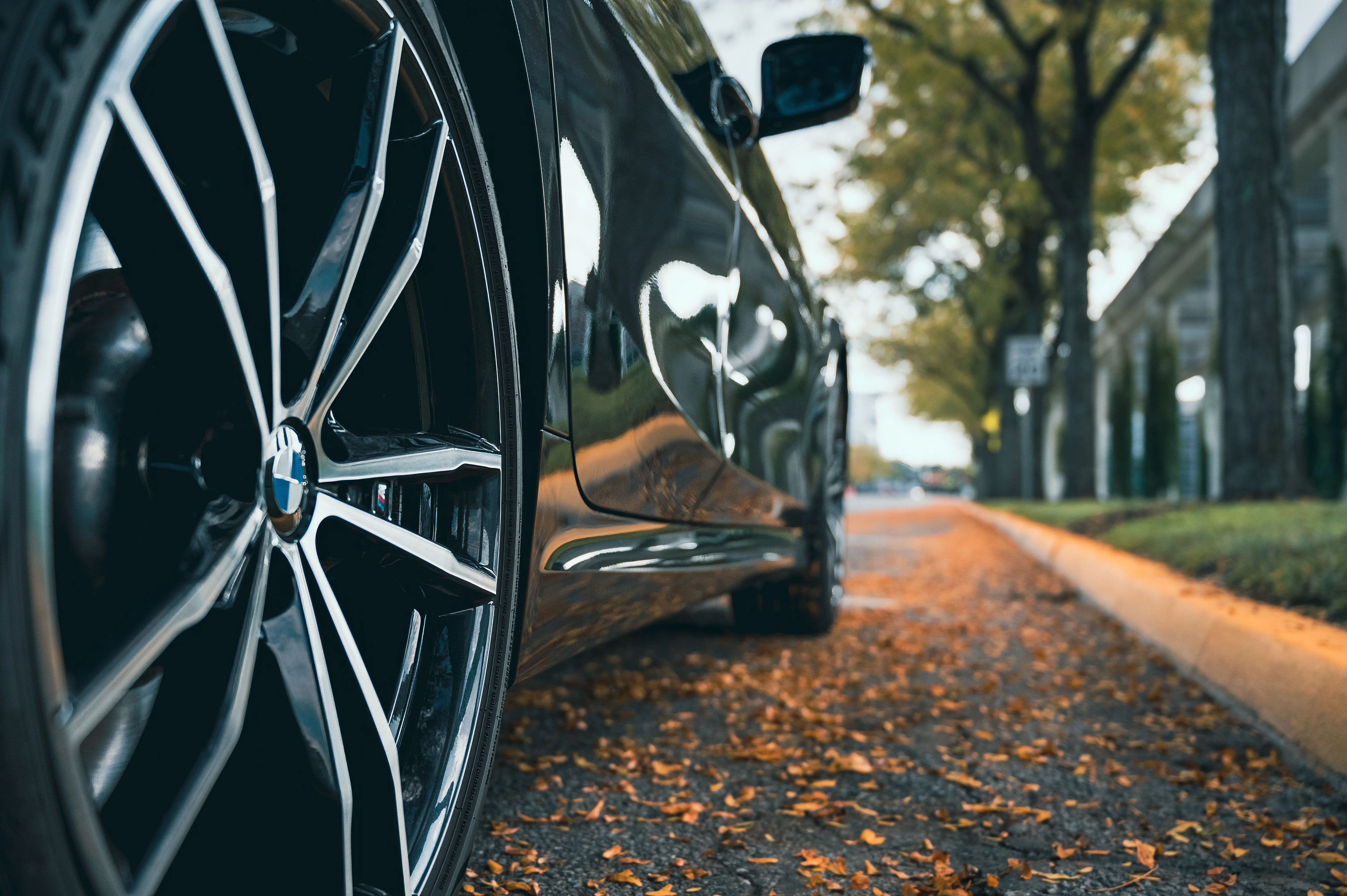 Close-up of a car's shiny alloy wheel parked on a tree-lined urban street with fallen leaves on the pavement.