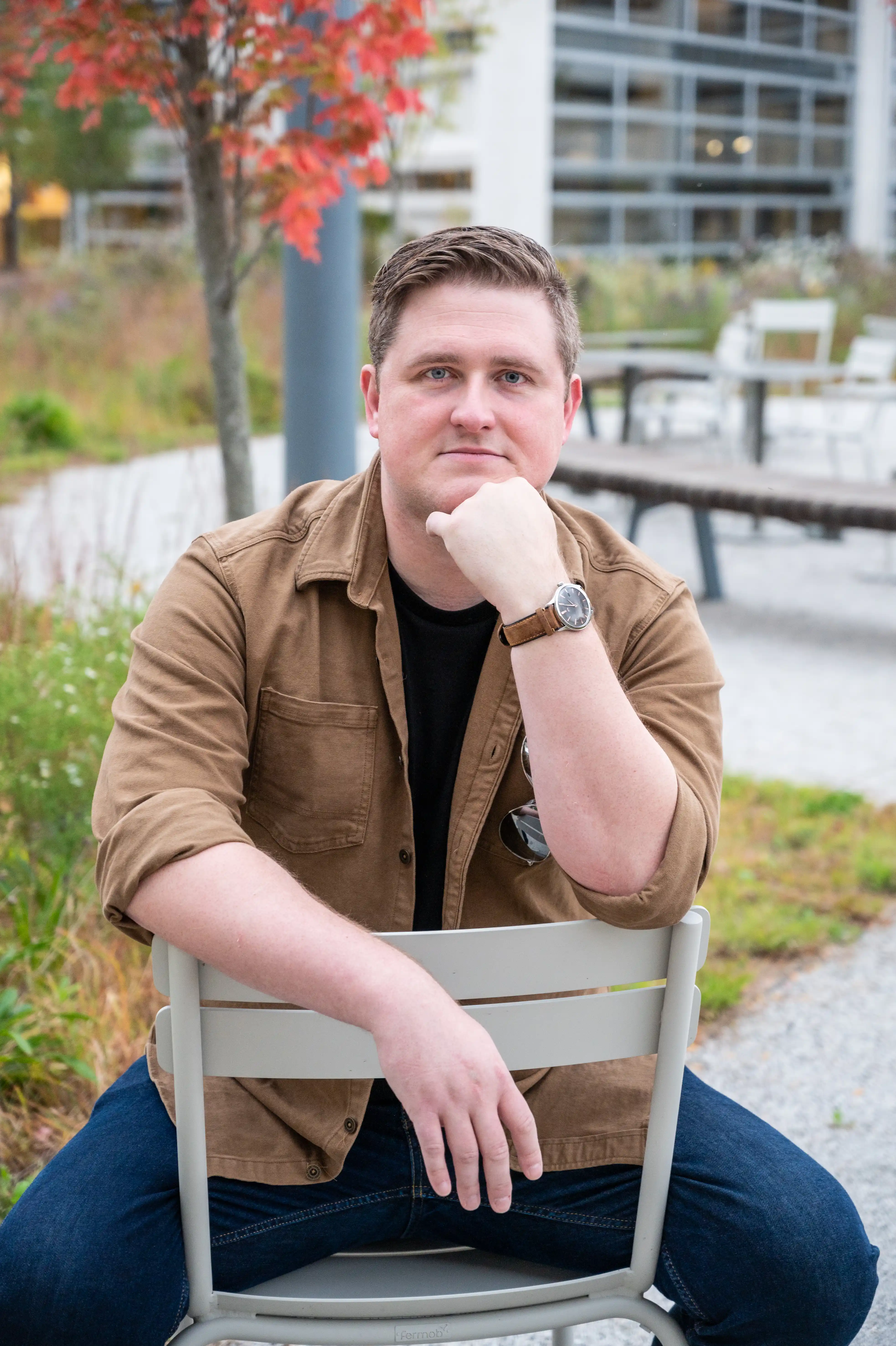 Man sitting on a metal chair outdoors with a thoughtful expression, wearing a brown jacket, black shirt, and blue jeans, with autumn foliage in the background.
