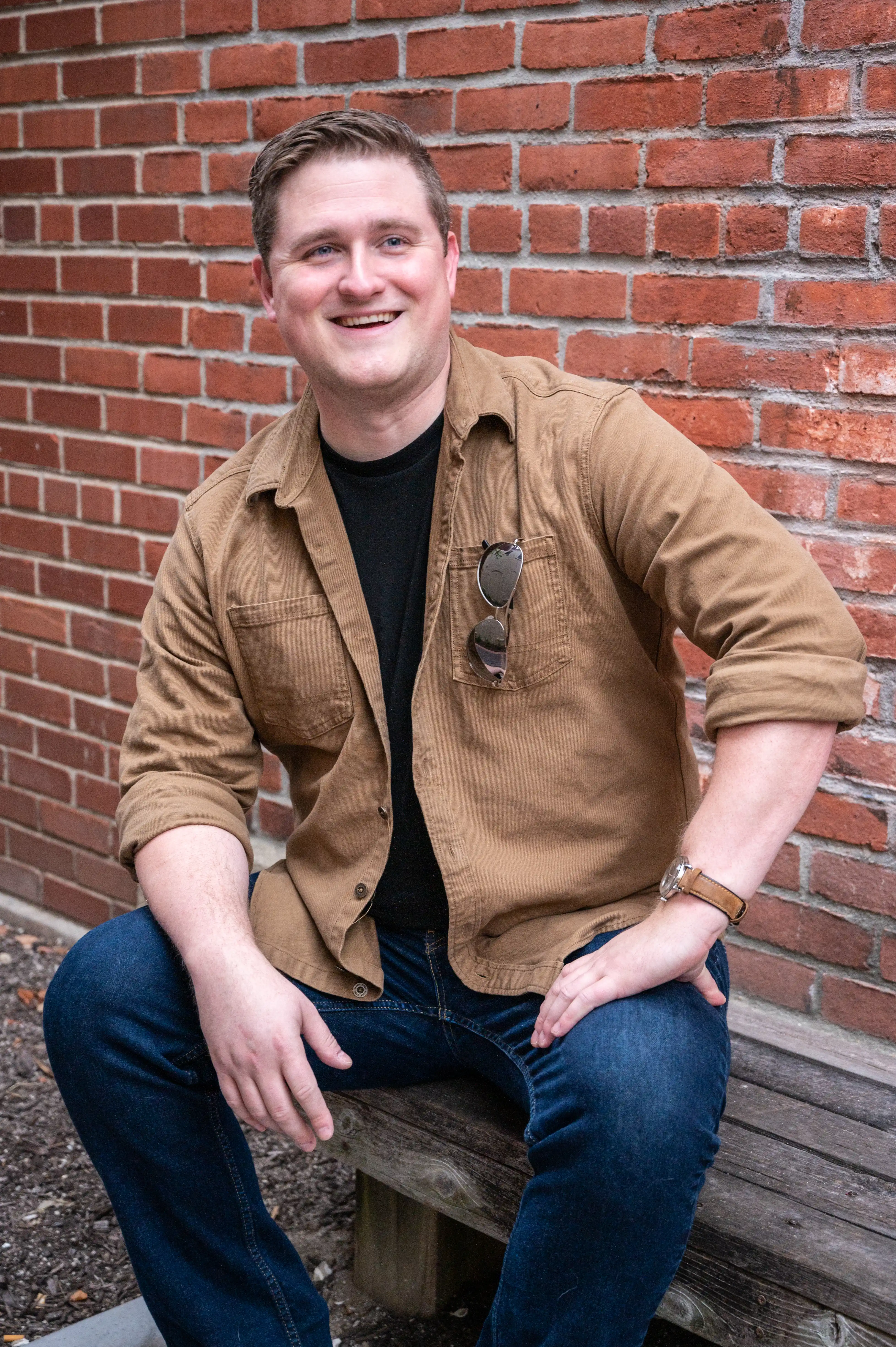 Man smiling at the camera while sitting on a step with a brick wall background.
