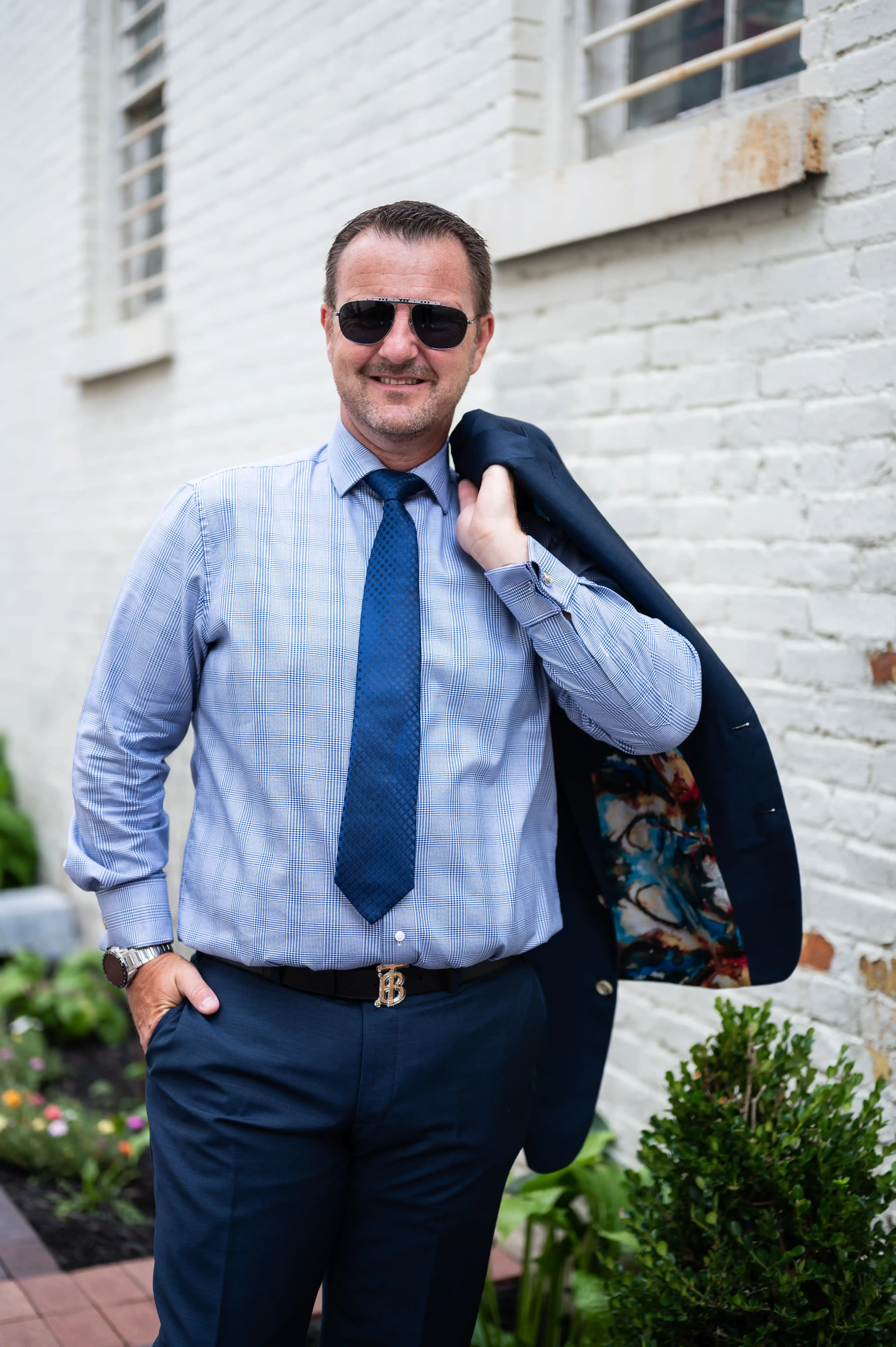 Confident businessman in a blue shirt and tie with sunglasses holding a jacket over his shoulder against a brick wall background.