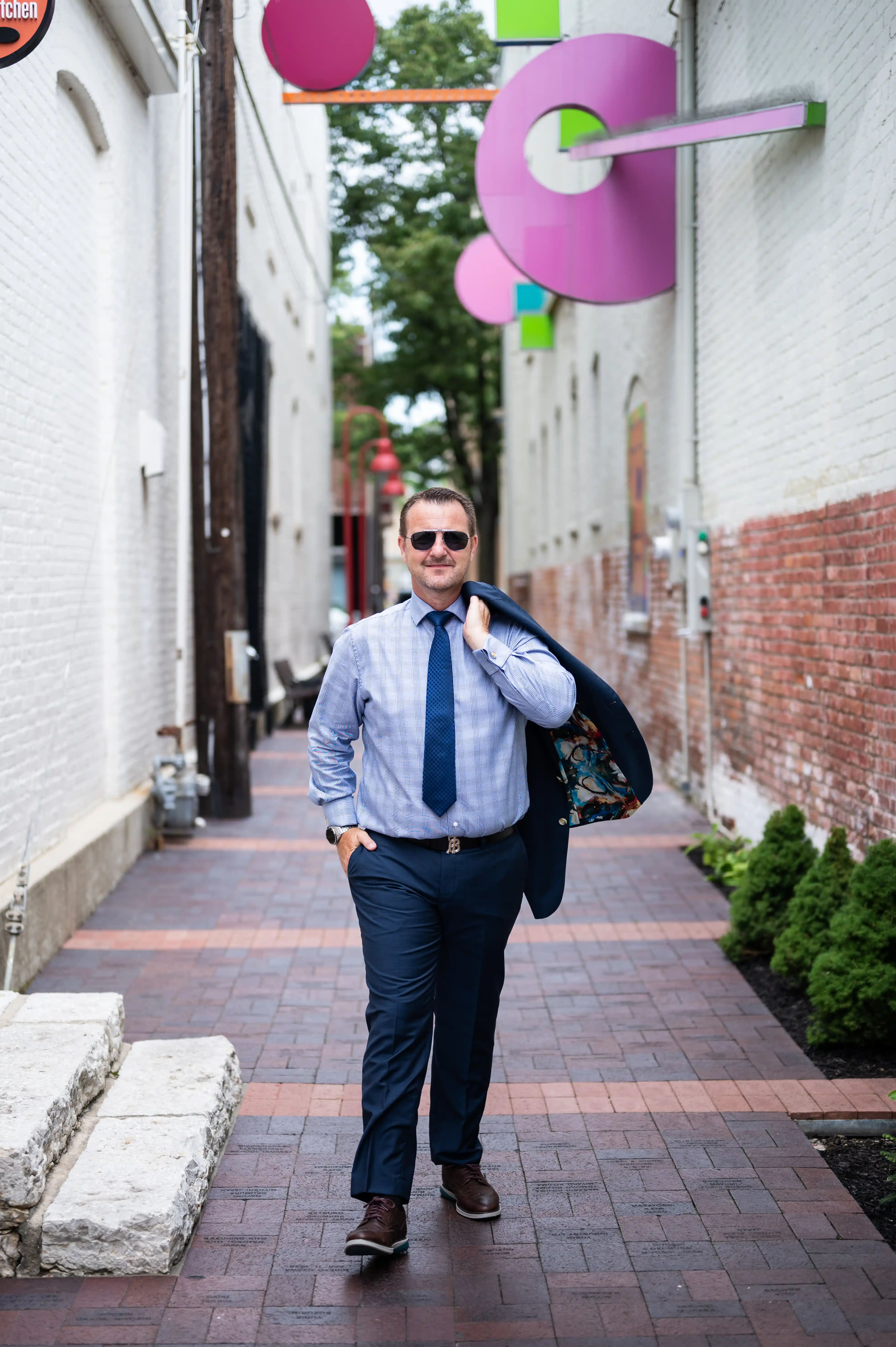 Man in a shirt and tie walking with sunglasses and a backpack in an alleyway decorated with colorful balloons.