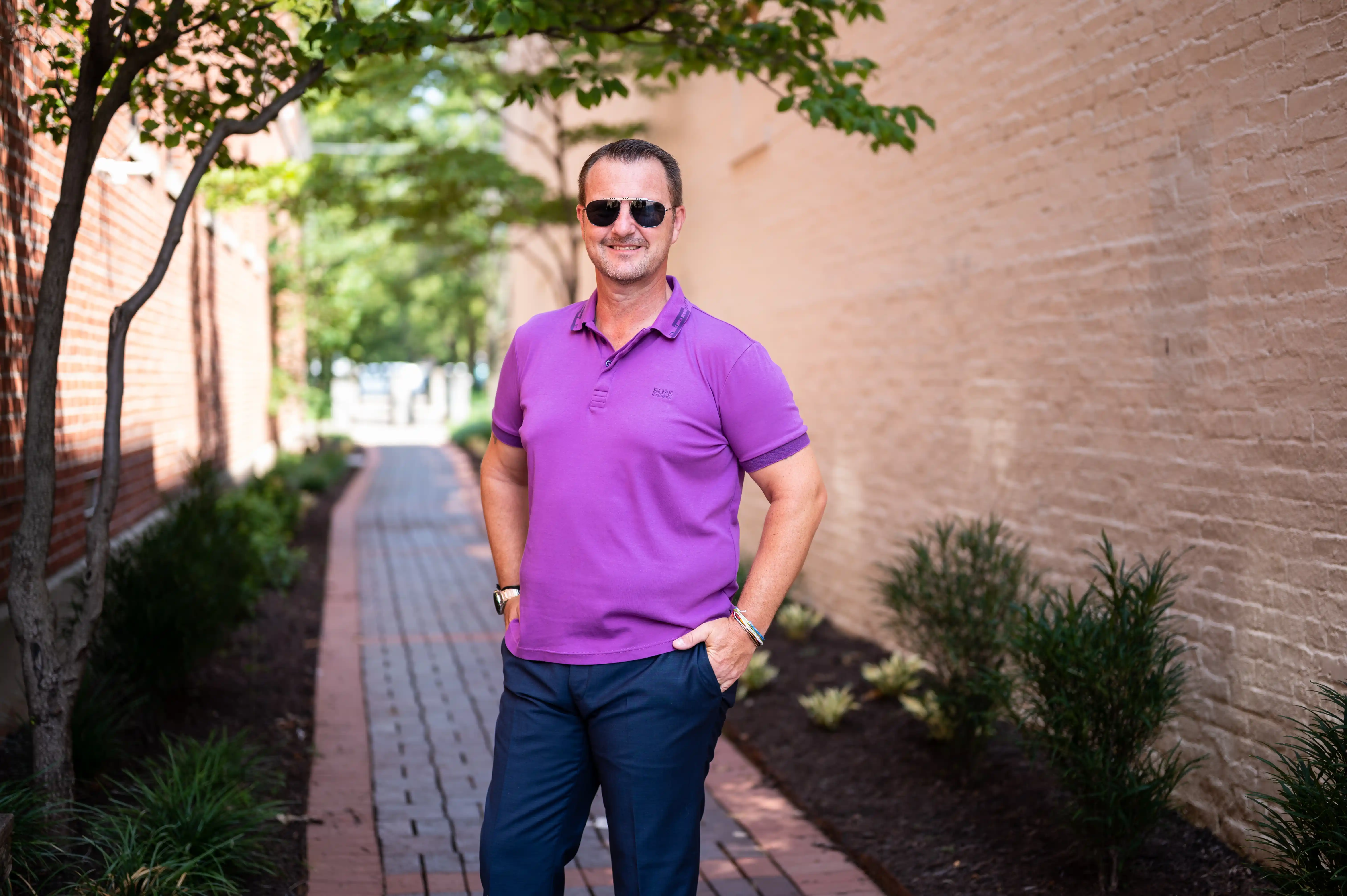 Man standing on a sidewalk with trees, wearing sunglasses and a purple polo shirt.