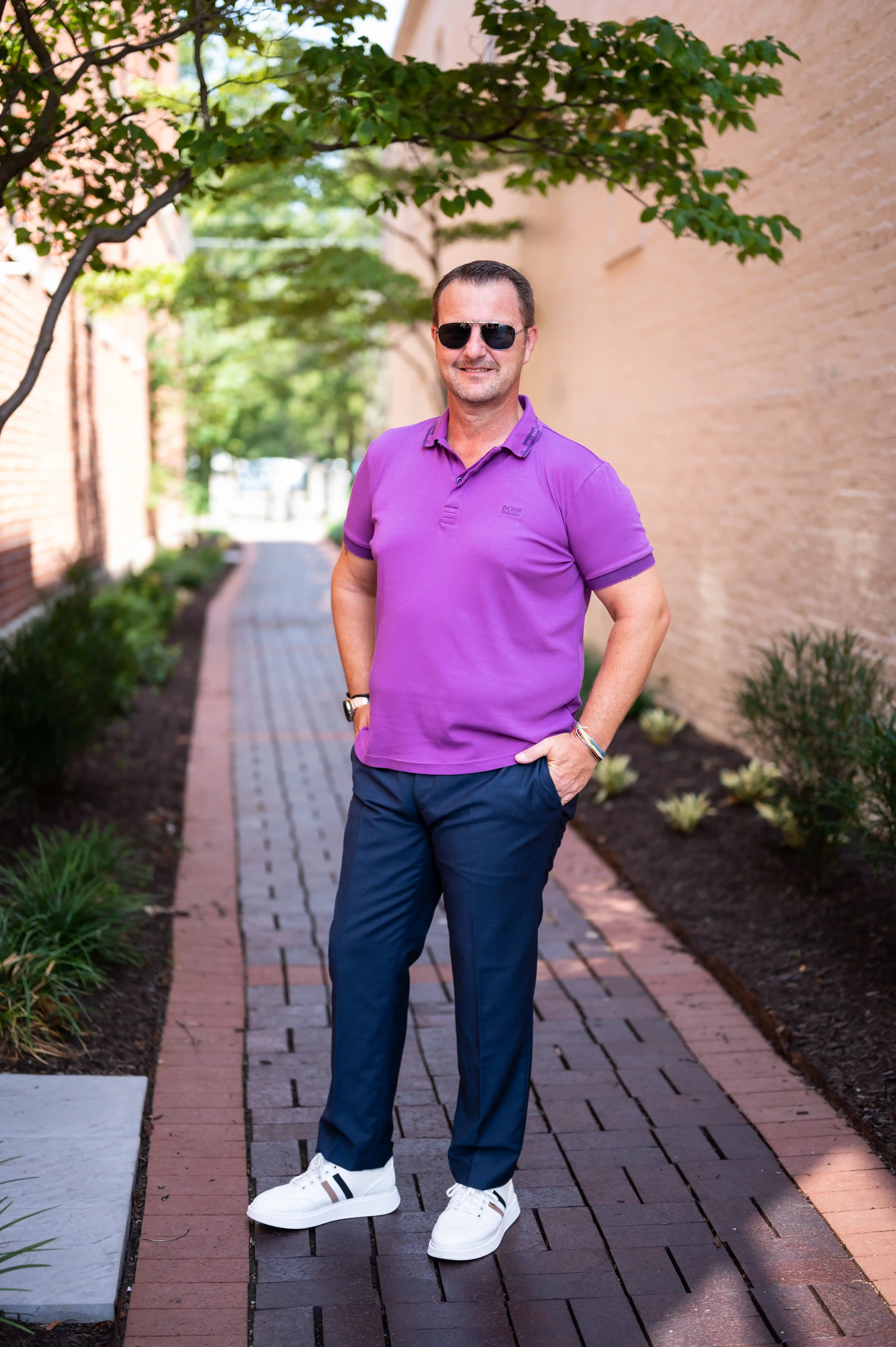 Man standing on a sidewalk wearing sunglasses, a purple polo shirt, blue jeans, and white shoes.