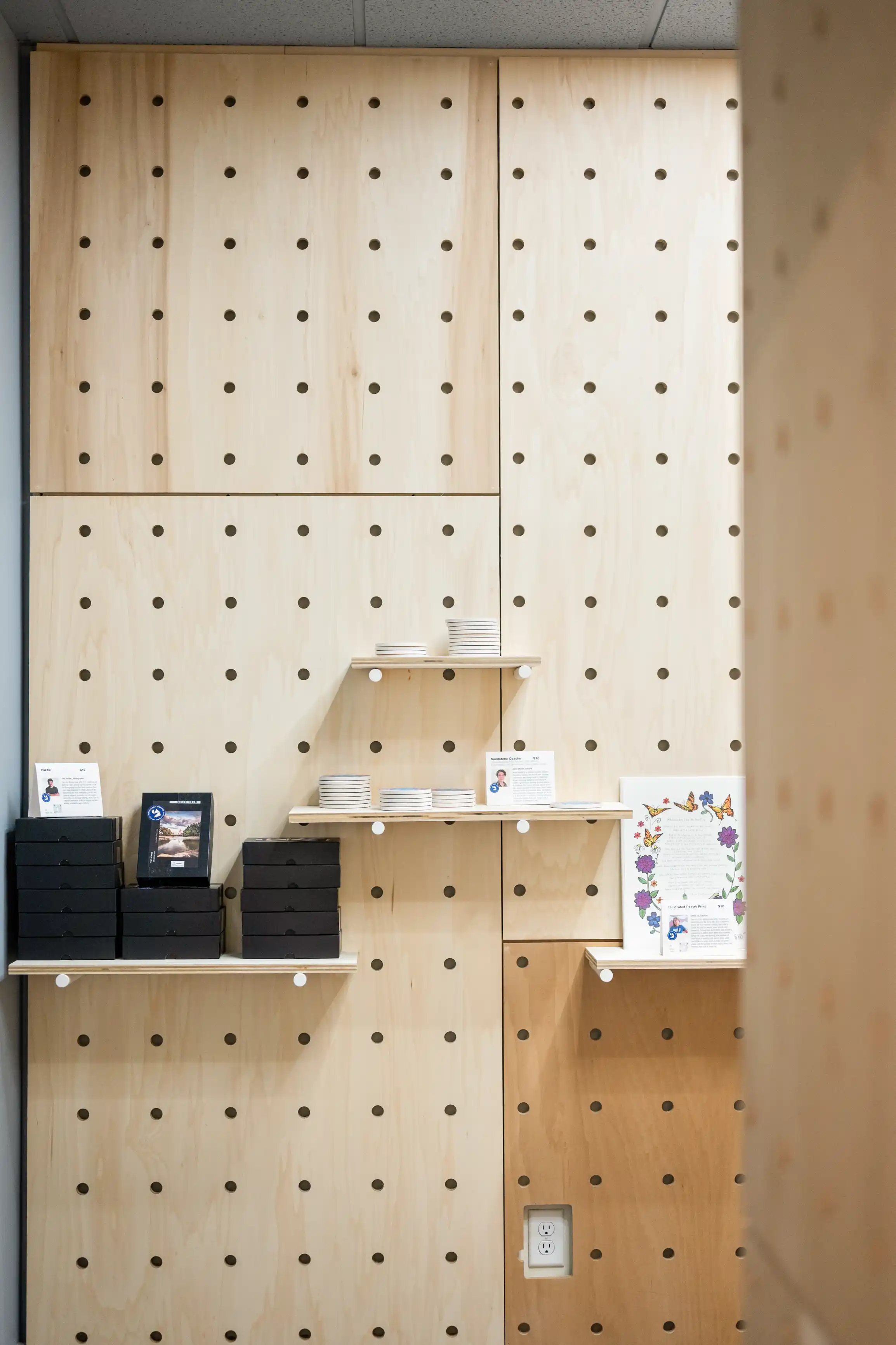 Interior view of a modern plywood wall shelving unit with circular cut-outs, displaying stacks of plates, black boxes, and promotional literature.