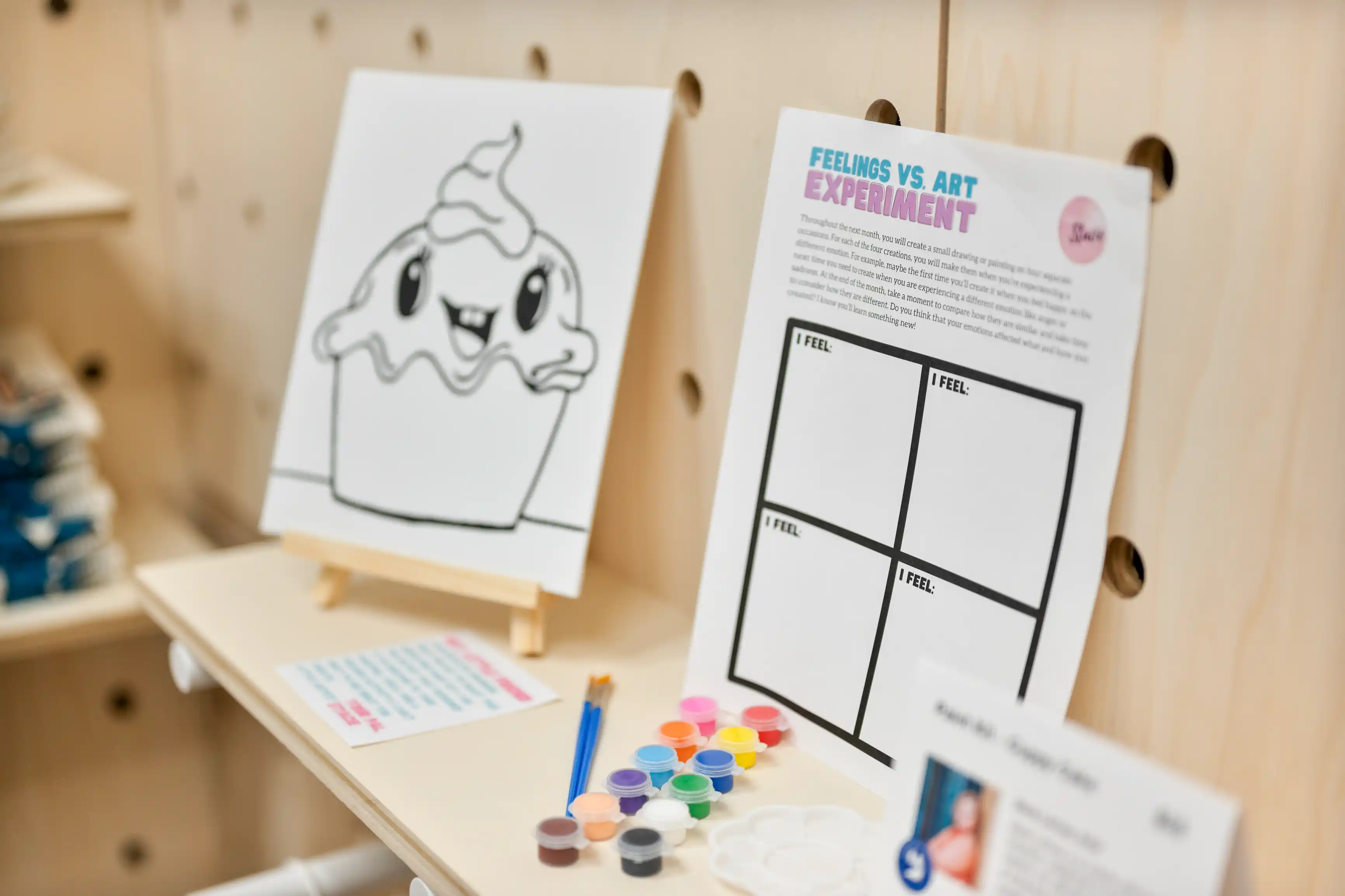 Art therapy station setup with a cartoon cupcake drawing on an easel, paint supplies, and an art therapy 'Feelings vs. Art' experiment worksheet.