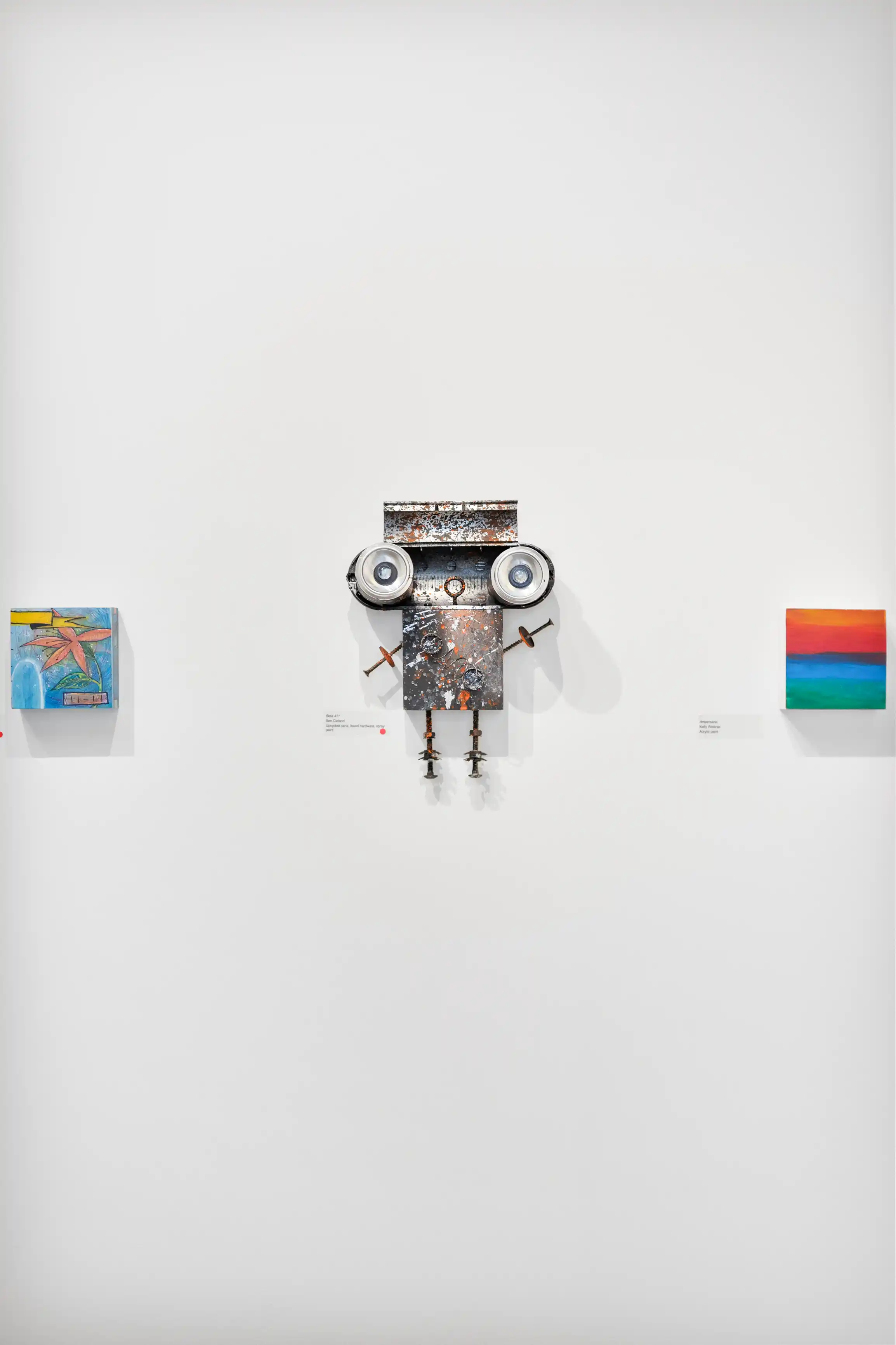 Mixed media artwork resembling a robot, surrounded by various paintings on a white gallery wall.