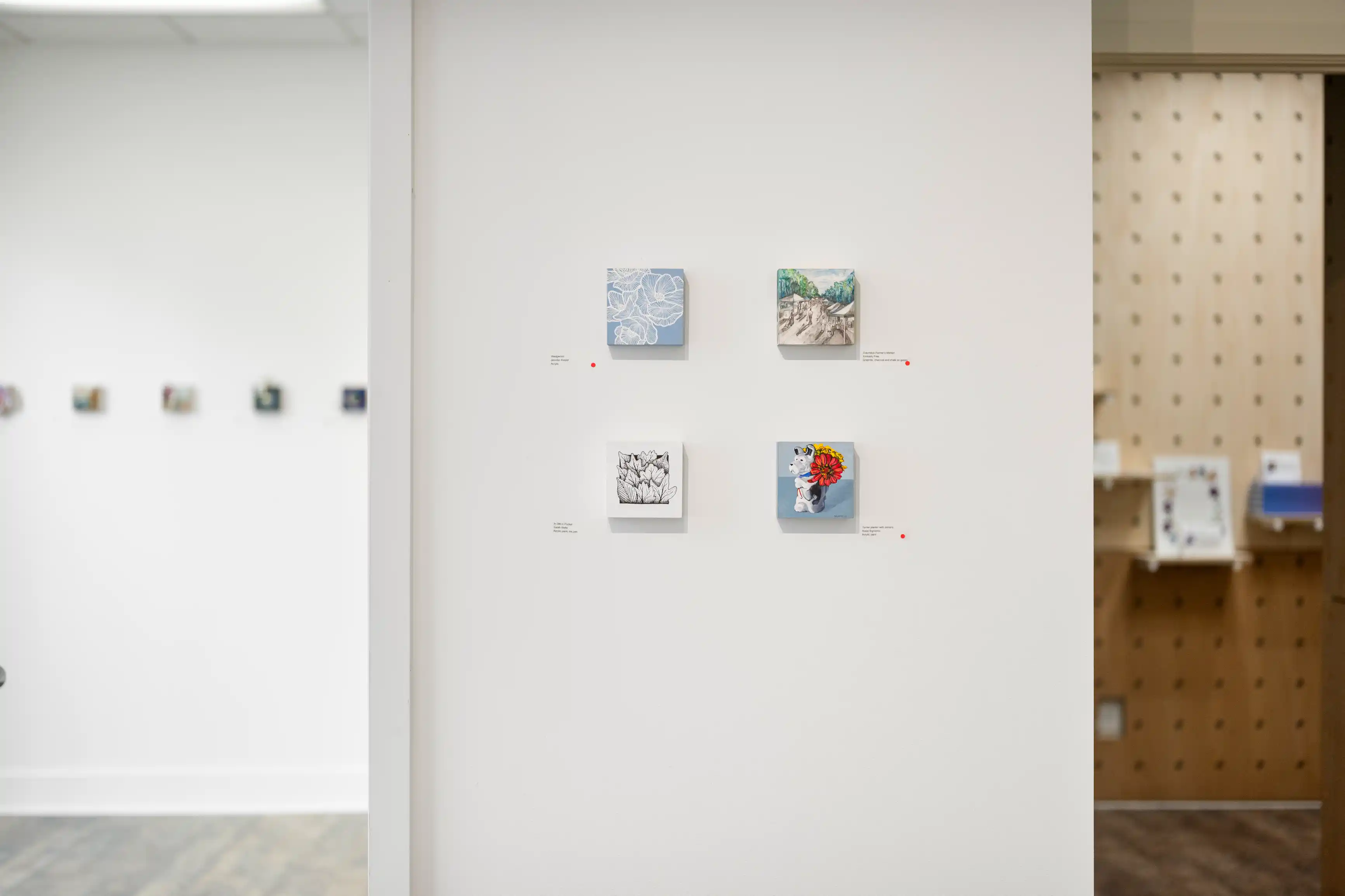 Art gallery interior with four small paintings displayed on a white wall, each accompanied by a descriptive label, and shelves with art materials in the background to the right.