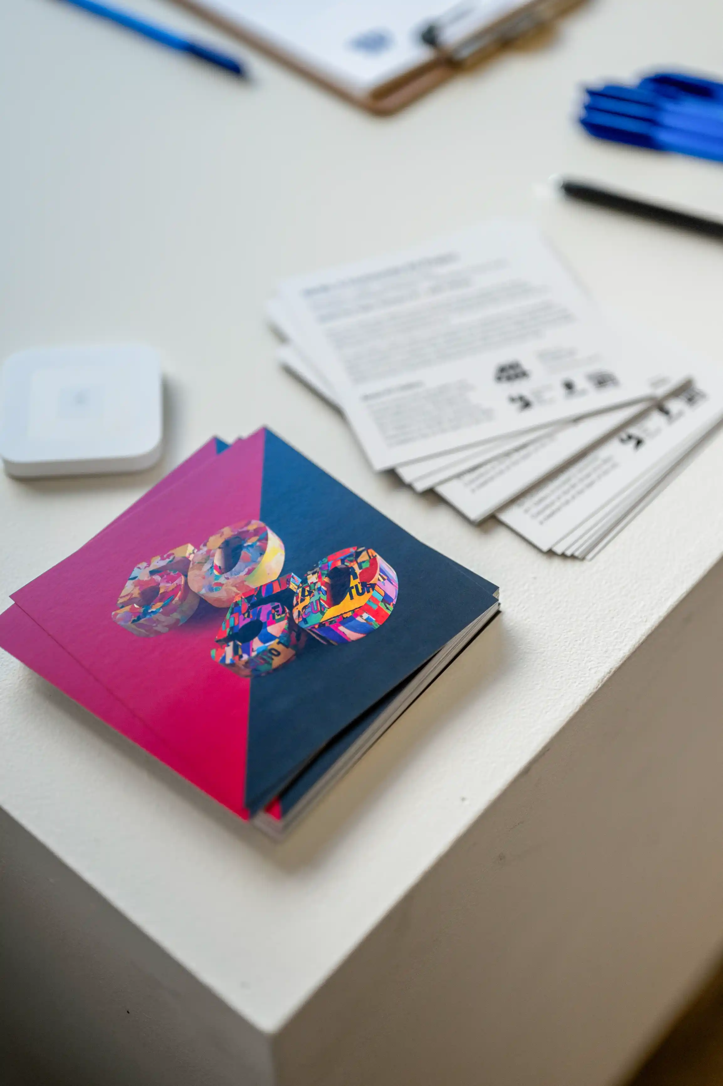 A stack of colorful brochures on a white table with documents and writing utensils in the background.