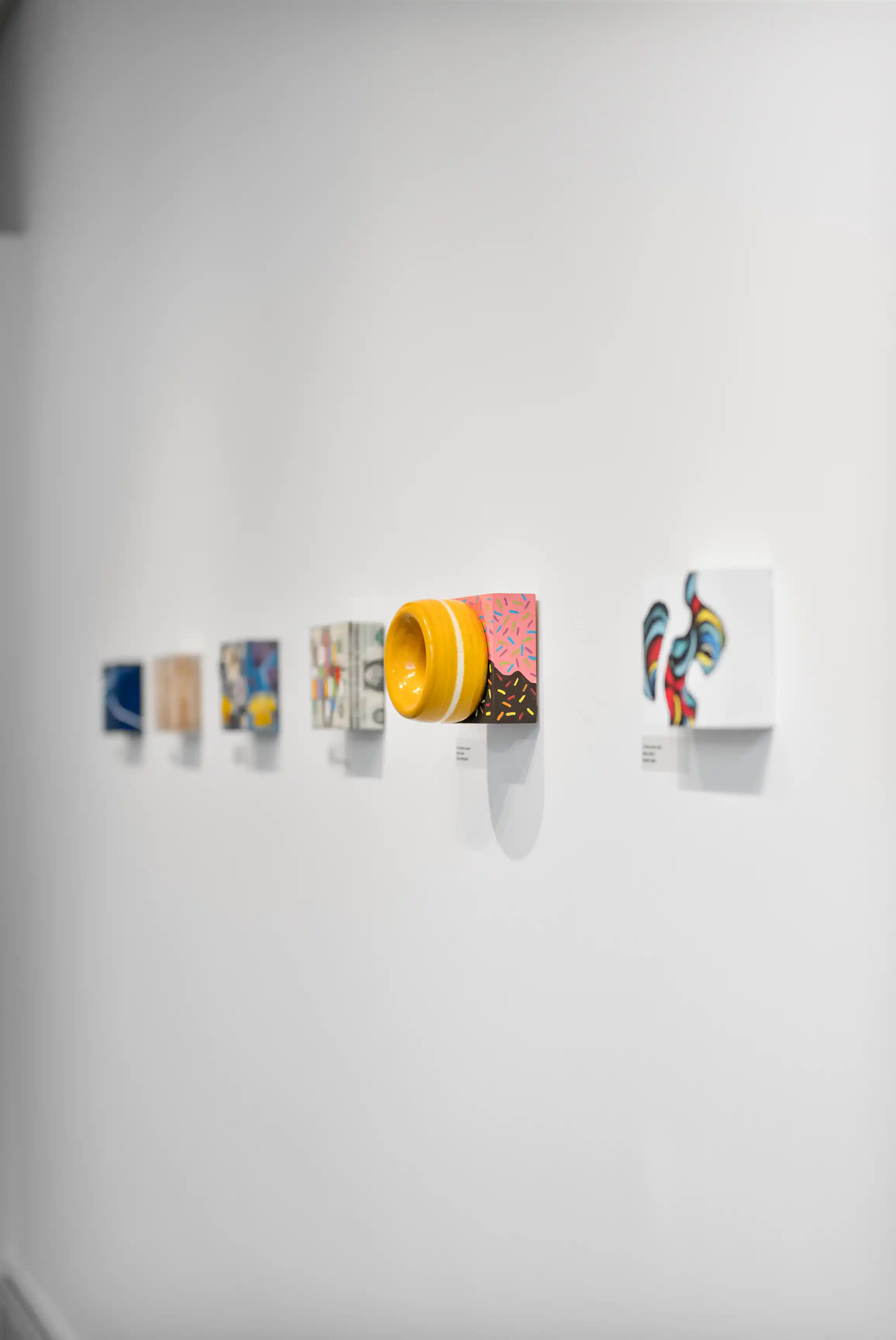 Contemporary art installation featuring a series of colorful abstract paintings and a yellow ceramic bowl displayed on white shelves against a white wall.