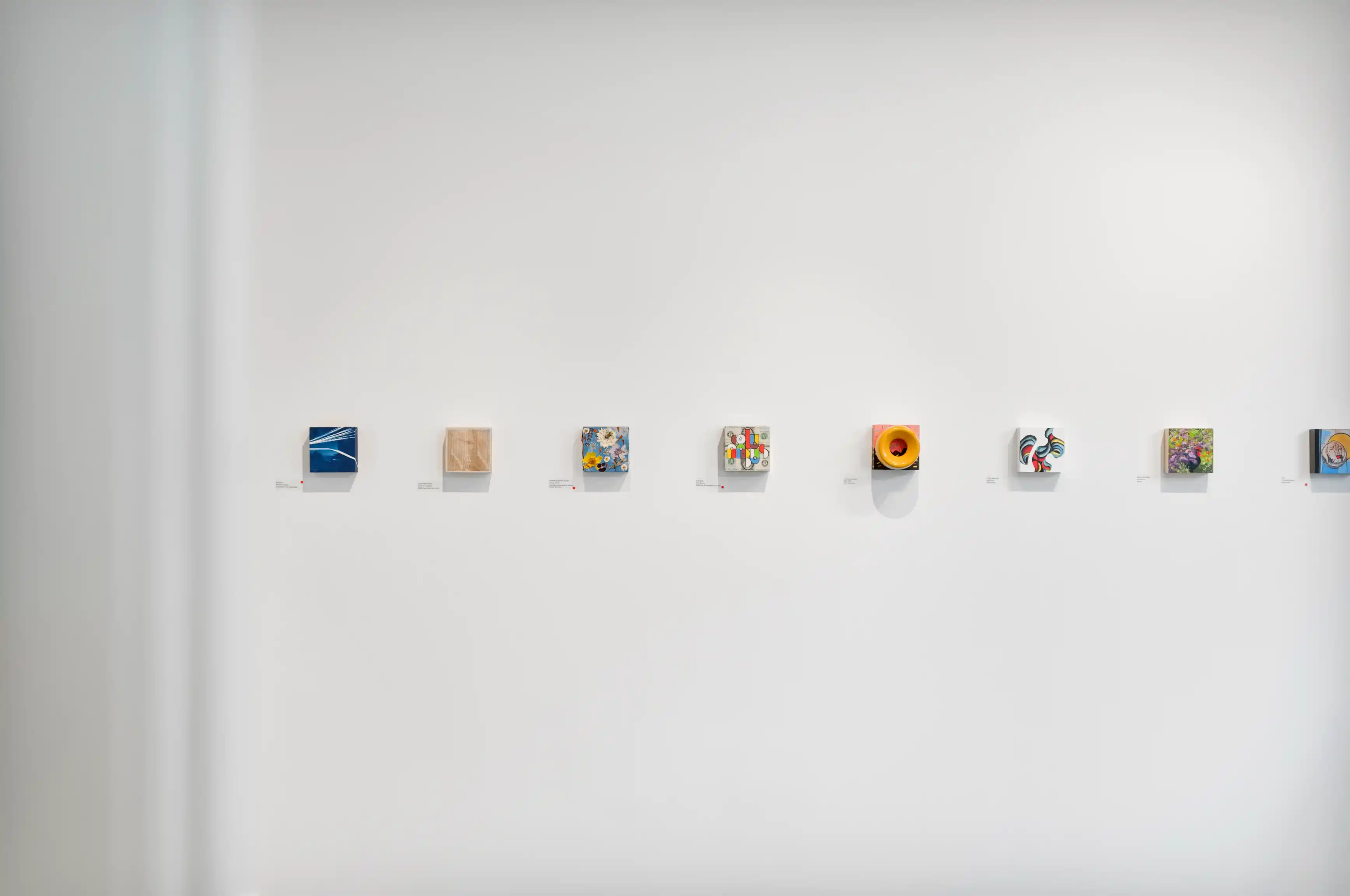 Art gallery wall showcasing a variety of small contemporary artworks with description labels underneath each piece.