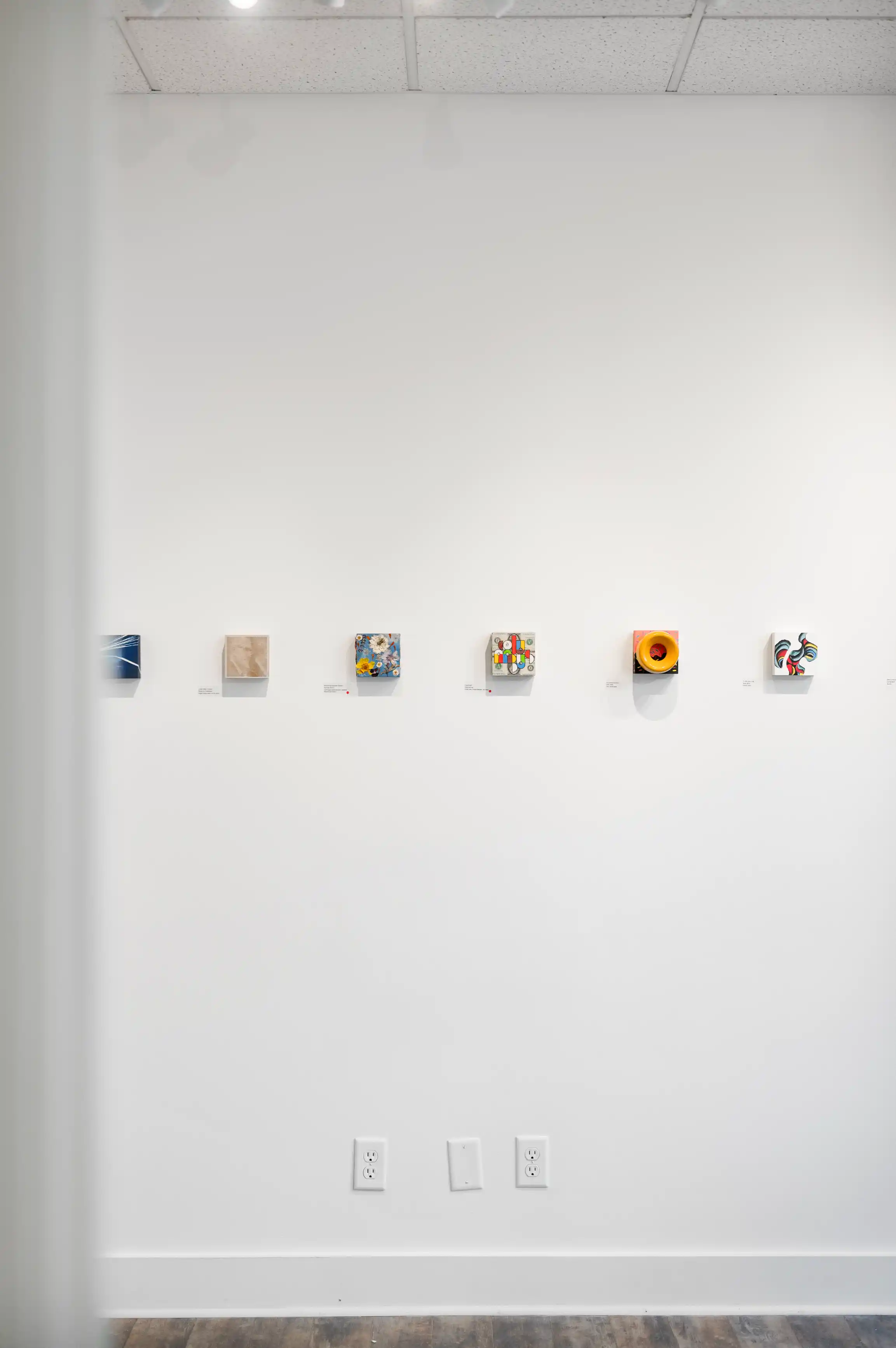 Art exhibition wall with a series of small, varied paintings lit by ceiling lights in a clean, white interior space.