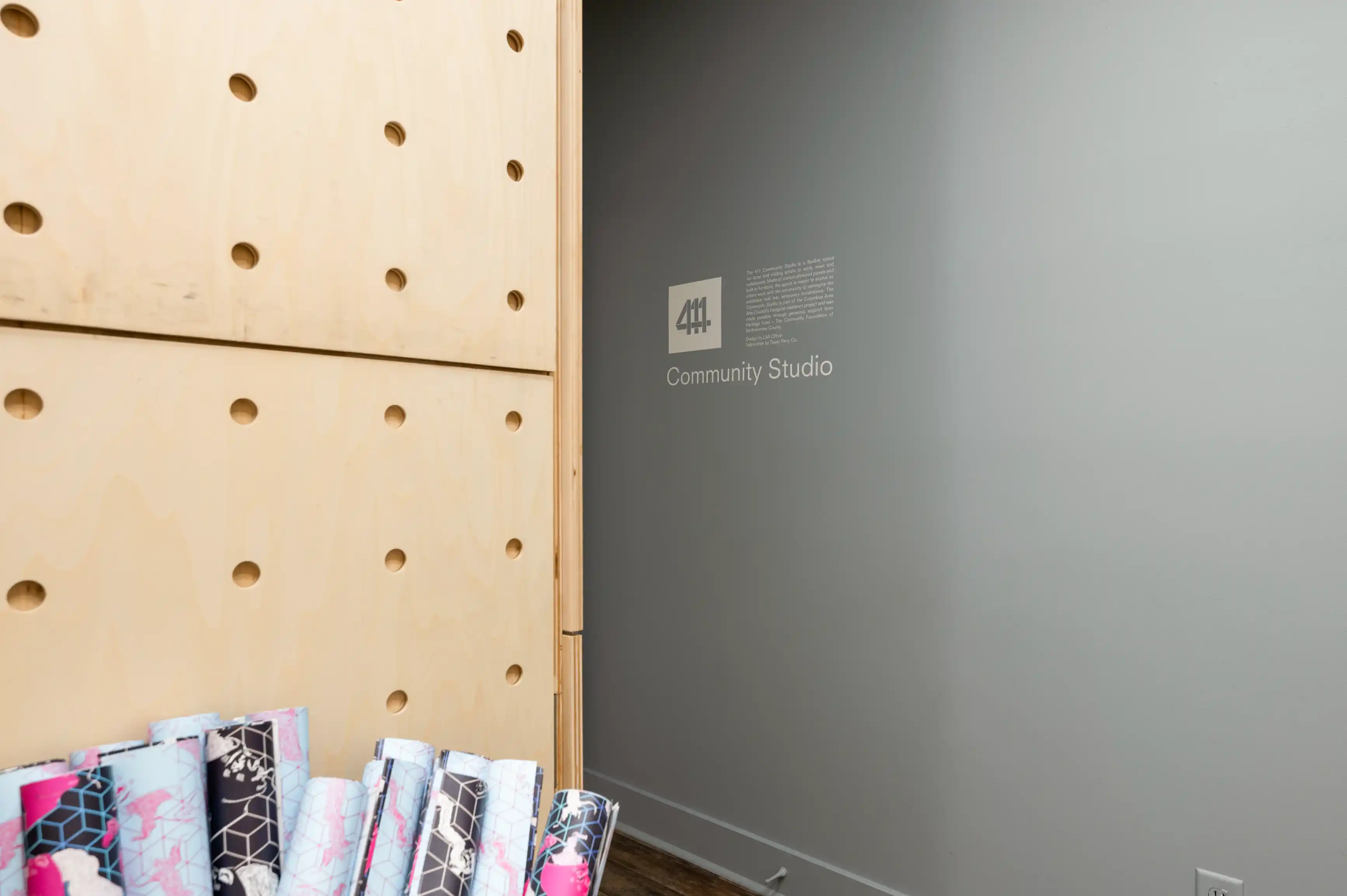 Corner of a modern room with a wooden panel with circular holes on the left and a wall with text and a sign reading "Community Studio" on the right, with a stack of colorful rolled yoga mats in the foreground.