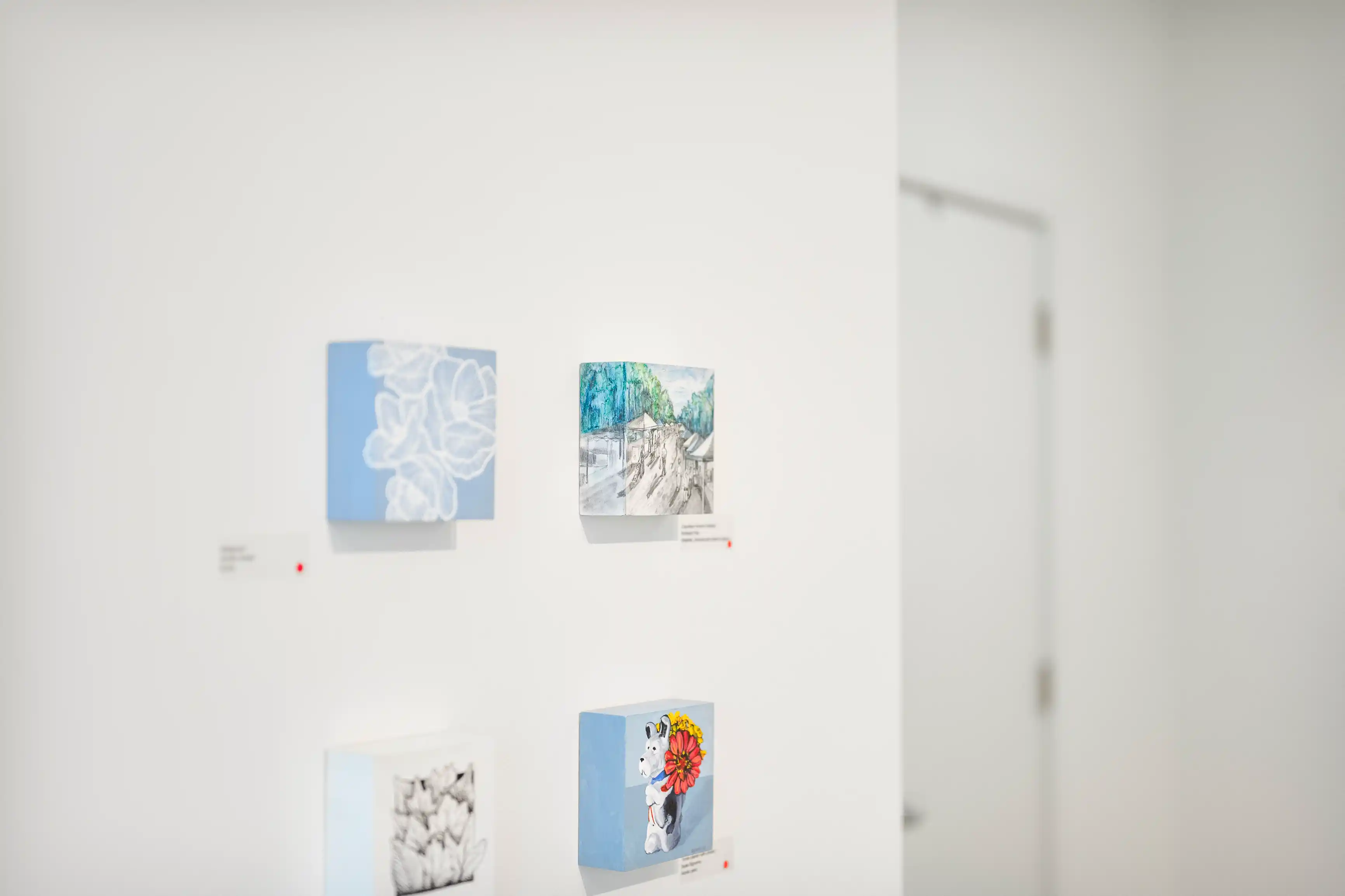 Three square canvases displayed on a white gallery wall, each with different artwork, with the focus on the vibrant painting of a cartoon dog holding a flower in its mouth.